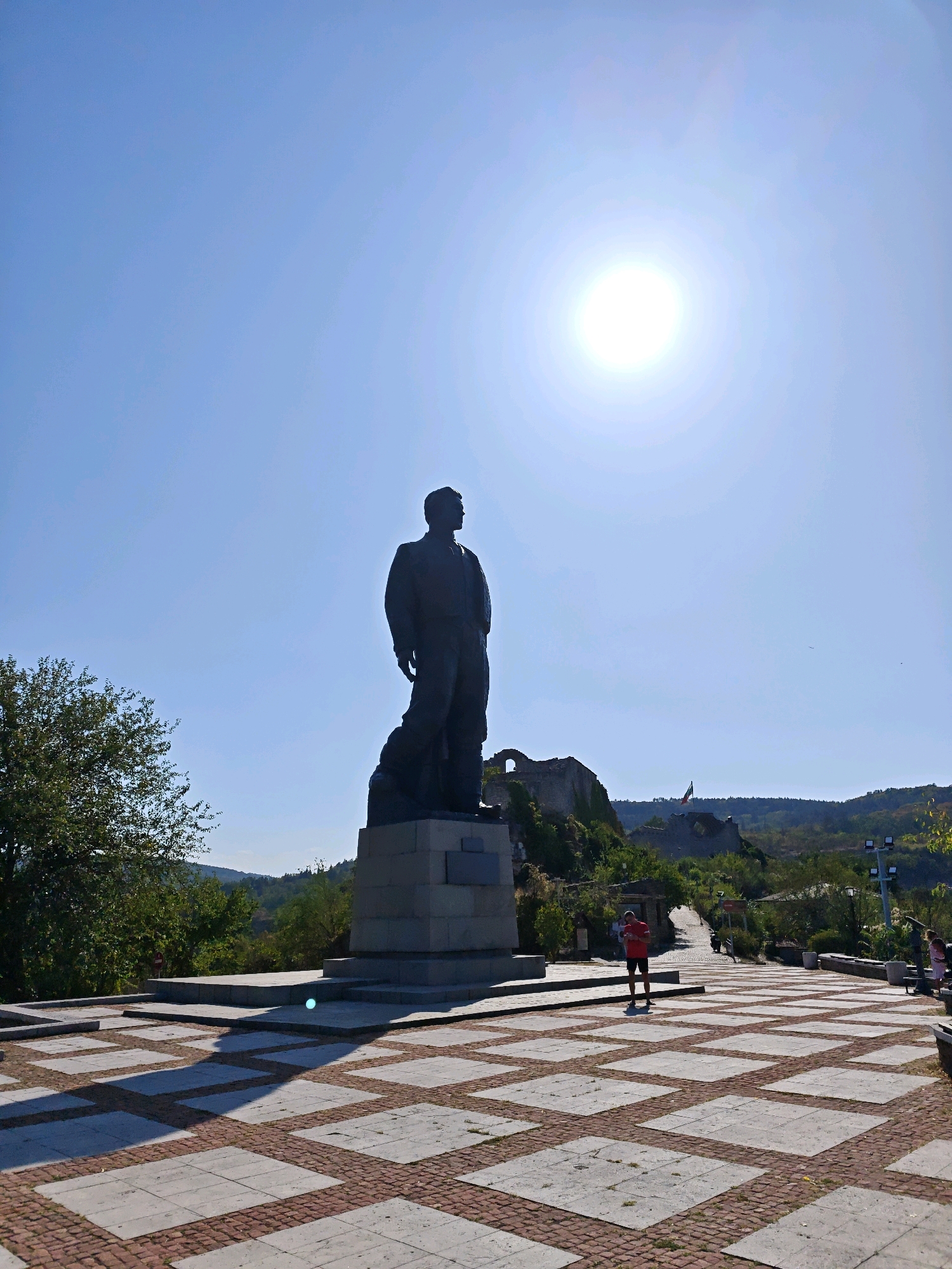 The image depicts the Monument to Vasil Levski in Lovech, Bulgaria, a prominent statue of the revolutionary figure standing tall against a clear blue sky. The sun is bright and positioned high in the sky, creating strong shadows on the paved plaza below. The plaza is tiled with a pattern of large square gray tiles and smaller red brick tiles.
In the foreground, the base of the statue is visible, constructed from large concrete blocks. The statue itself is a dark silhouette of Levski, looking towards the horizon, with a cape draped over his shoulder.
To the right of the statue, a person wearing a red t-shirt and dark shorts is standing, looking down at a device in their hands. Further in the distance, a section of a castle ruin is visible on a hill, with a Bulgarian flag flying. The surrounding landscape is hilly and covered with green trees. The overall scene is bright and sunny, with a sense of historical significance and public space.
