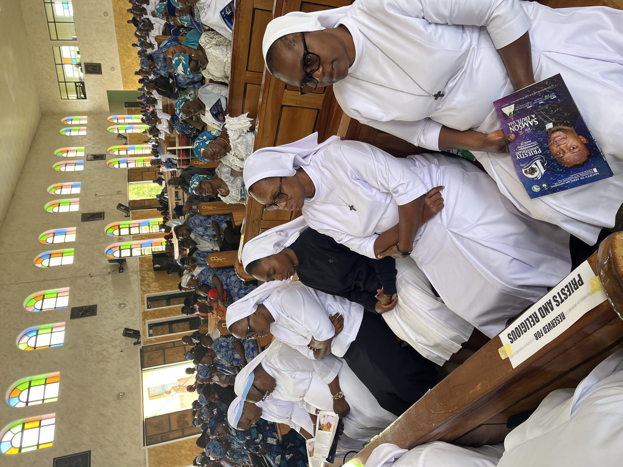 The image depicts a group of people seated in what appears to be a church or religious institution, possibly in Vom, Nigeria. In the foreground, several women in white religious habits are seated on wooden pews. One woman on the far right is holding a booklet with an image of a man and text that appears to be in English. The women are wearing glasses and appear to be attentive. The pews are marked with a sign that reads "RESERVED FOR PRIESTS AND RELIGIOUS." In the background, more people are seated, many wearing blue and white patterned clothing, suggesting a congregation. Stained-glass windows with colorful designs are visible along the upper walls of the building. The overall atmosphere appears to be one of a religious gathering.