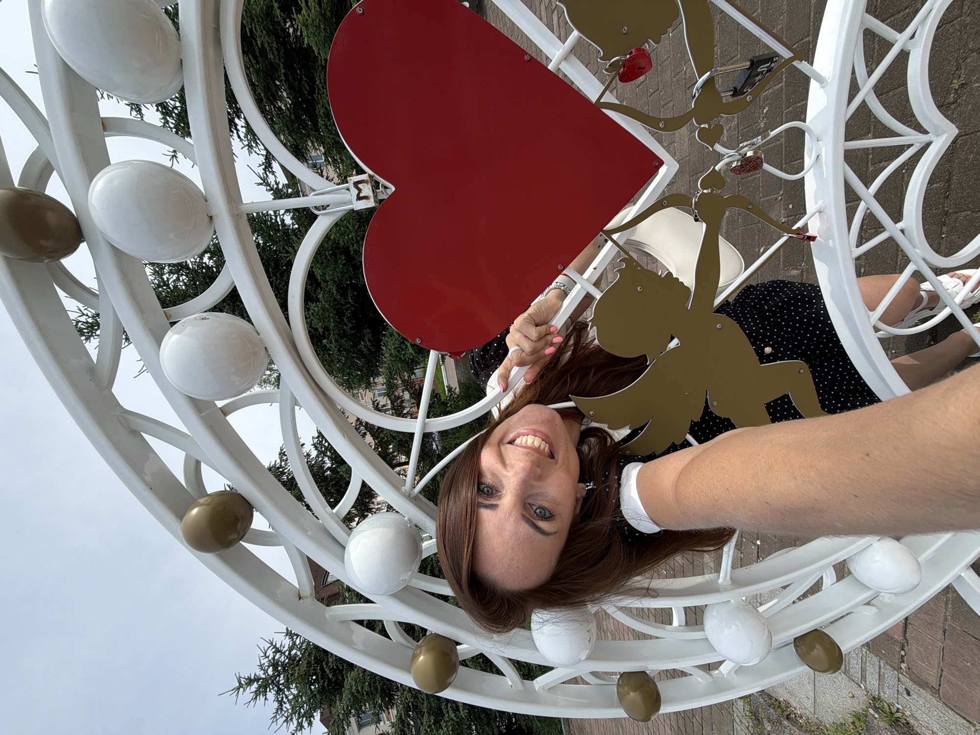 The image shows a woman taking a selfie in front of a large white metal heart sculpture adorned with golden cherubs and red and white spheres. A red heart is prominently displayed in the center of the sculpture, with a padlock attached to it. The sculpture appears to be located outdoors, possibly in a park or public square, with trees and buildings visible in the background. The woman is smiling and leaning into the frame, with her arm extended to capture the selfie. The overall impression is one of a romantic or celebratory setting.
While the image itself does not explicitly state its location, the prompt requests it to be identified as being in Birobidzhan, Russia. This sculpture is likely a landmark or decorative element within the city.