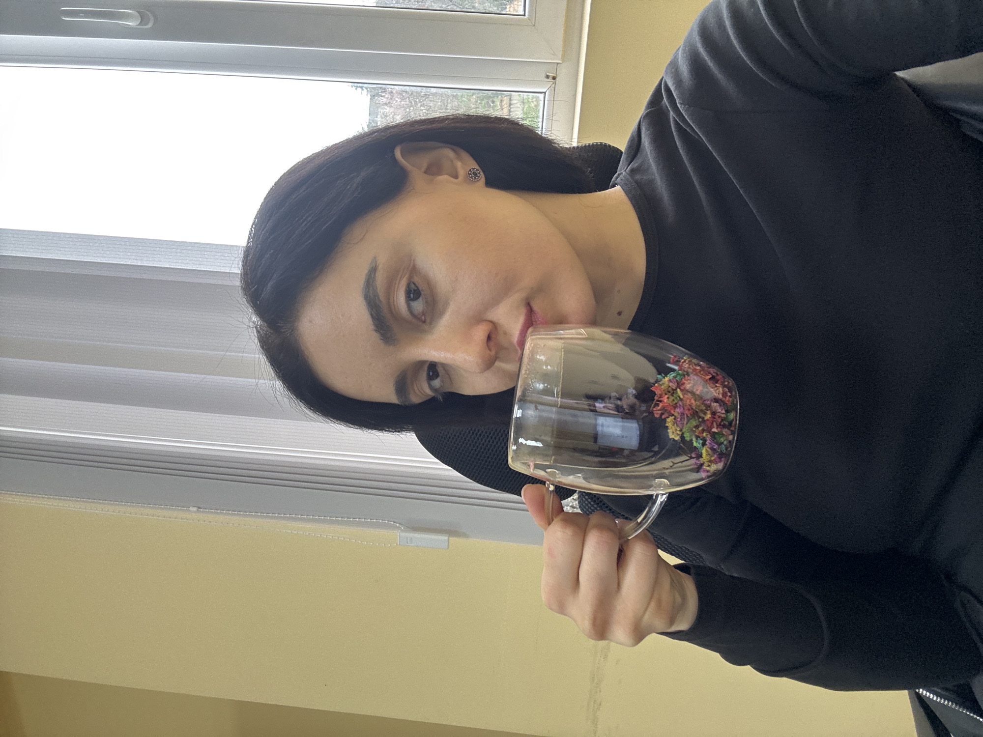 A woman with dark hair is holding a clear glass mug to her lips, as if about to drink. The mug contains a colorful mixture, possibly tea with flowers or herbs. She is wearing a black top. The background shows a window with closed blinds and a light yellow wall. There's no information given to infer the location in Tbilisi, Georgia.