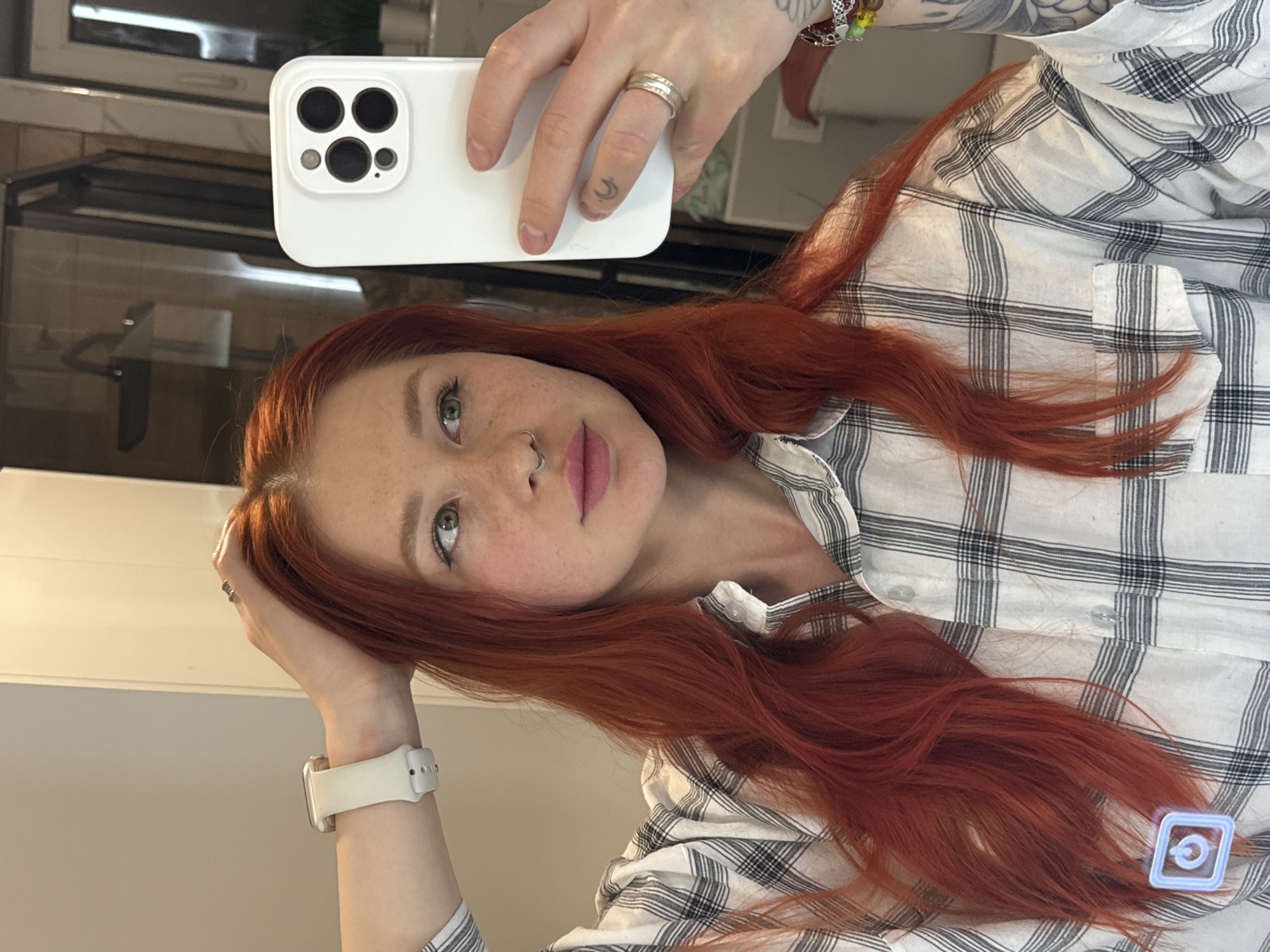 A woman with red hair and freckles is taking a selfie with a white smartphone. She wears a white and black plaid shirt and a white smartwatch. She is wearing a silver ring on her right hand's ring finger. Her hair is long and wavy, and her makeup appears minimal. The background includes a reflection in a mirror, showing a window and what appears to be a kitchen setting with a stainless steel oven. The location or mention of the city of Rostov-on-Don, Russia, is not discernible from the image.