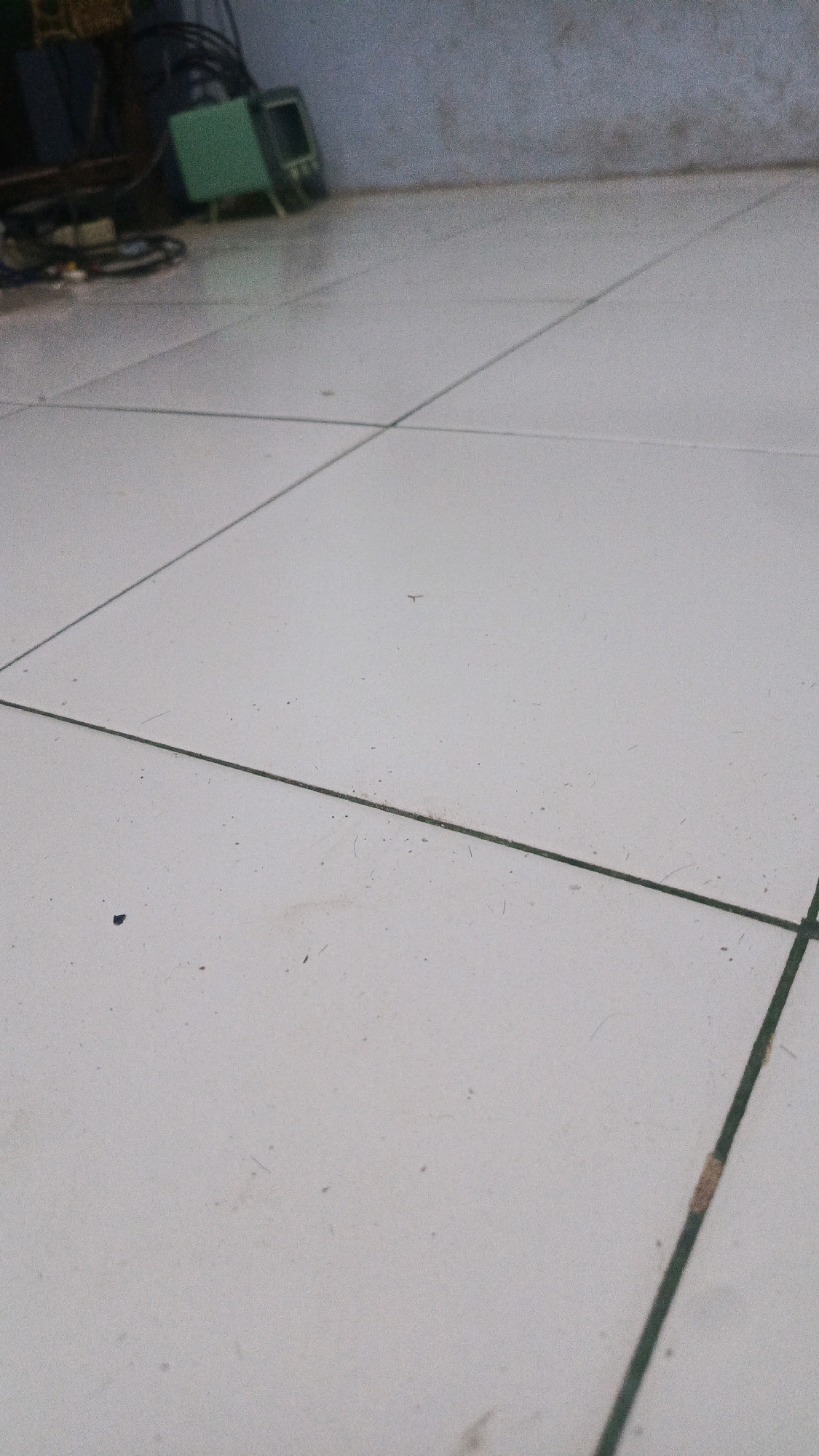 The image shows a tiled floor with a low-angle perspective. In the upper left corner, there are some blurry objects, including a green electronic device with a screen and a bundle of cables. The floor is light-colored with visible grout lines forming a grid pattern. Some small dark specks, possibly dust or debris, are scattered across the tiles. The background is a textured wall, also out of focus. The overall scene appears to be indoors, likely in an unfinished or utility area. There are no people or any discernible activities taking place.