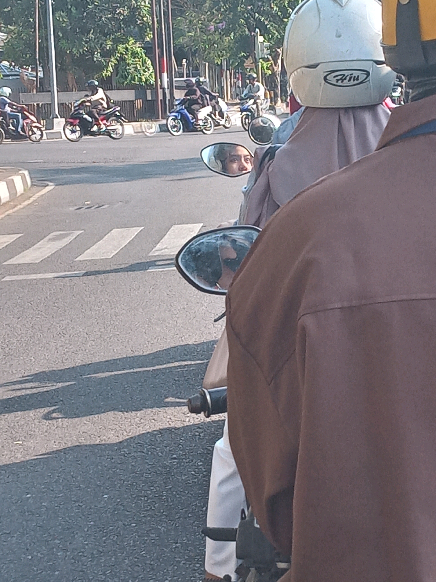 In Yogyakarta, Indonesia, a brown-clad individual is riding on the back of a motorcycle, partially obscuring the view. In front of them, a person wearing a white helmet and a light purple hijab is also on the motorcycle. A rearview mirror reflects the face of the person in front, showing a young woman with dark hair and noticeable eyebrows. Other motorcycles are visible on the road, some carrying passengers. The scene takes place on a paved street with a pedestrian crossing marked by white stripes. Trees and utility poles line the street, suggesting an urban or suburban environment. The lighting indicates it is daytime, with shadows cast on the road.