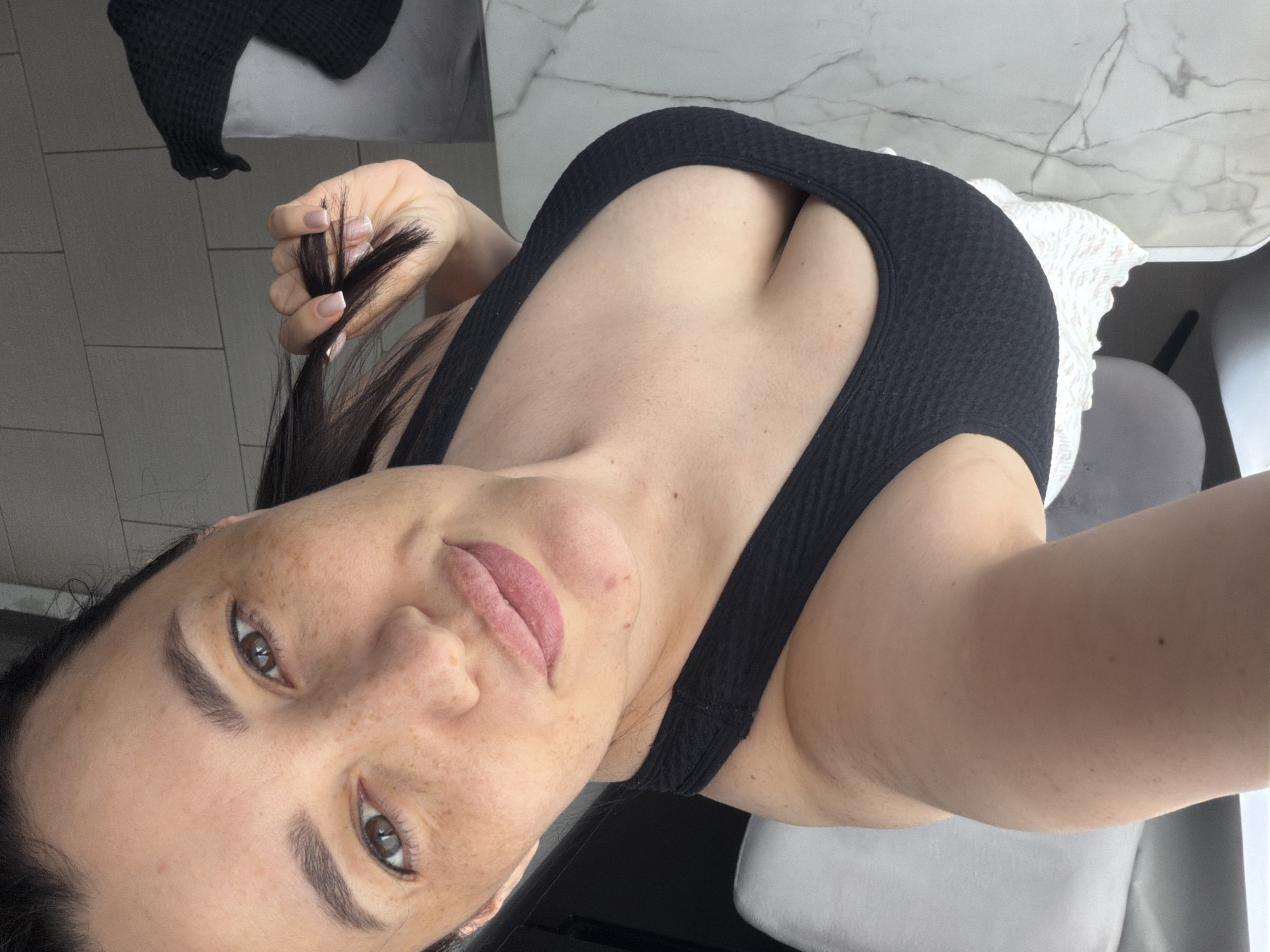 A female individual is depicted in a close-up, high-angle selfie. She has dark hair, dark eyes, a light skin tone with numerous visible freckles across her face, and full, slightly pursed lips. Her right hand, exhibiting a French manicure, holds strands of her dark hair near her head. She wears a black, textured or ribbed sports bra/top, revealing cleavage, and a white garment with a ruffled edge below. In the background, to the left, a portion of a white chair with a grey backrest is visible. To the right, a marble-patterned countertop or table surface extends, beside which a dark, knitted fabric object rests on a white surface, possibly bedding or another chair. Above the dark object, a light-colored tiled wall is partially visible. The scene is an indoor setting within Водоканал Снт, Russia.