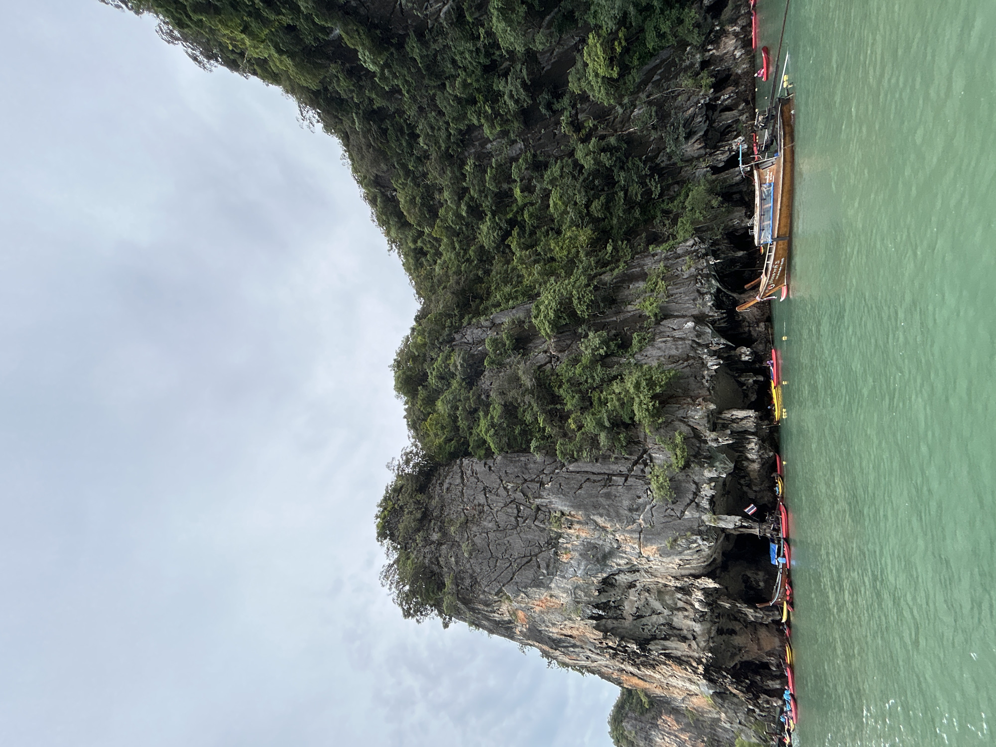 In Krasom, Thailand, a longtail boat is anchored in emerald green water near a sheer, towering cliff face. The cliff is covered in lush green foliage, with exposed rock formations showing cracks and striations. Several red, yellow, and other colorful kayaks are lined up near the base of the cliff, some occupied by people. The sky is overcast with soft gray clouds.