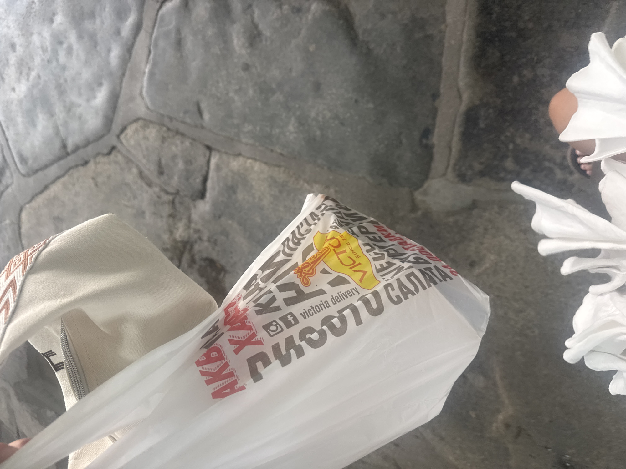 A person holds a white plastic bag with red and gray lettering that reads "AKBA XAP" and other unreadable text, as well as a logo for "VICTORIA delivery SINCE 1994." The bag is partially obscuring a beige fabric tote bag with a patterned band. The background consists of large, uneven gray stone paving slabs. The scene is likely outdoors, given the paving and natural lighting. There is no indication of the city of Plovdiv, Bulgaria.