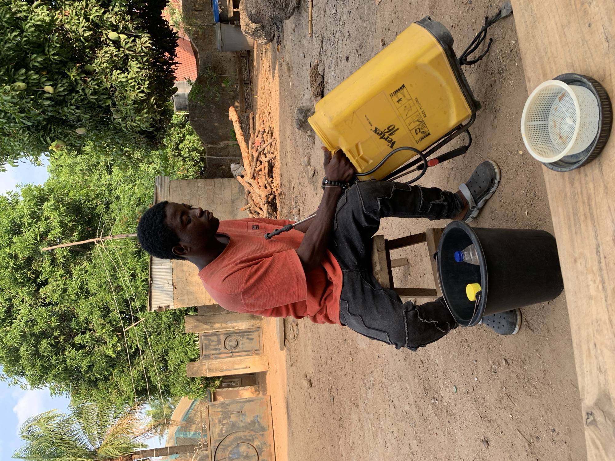 A man is seated outside in Akwanga, Nigeria. He is wearing an orange t-shirt, dark jeans, and dark Crocs. He is seated on a wooden stool. He is pouring liquid from a yellow Vertex 16 liter backpack sprayer into a black bucket that contains two small bottles with yellow and blue caps. The cap for the sprayer rests on a piece of wood nearby. Behind him is a pile of branches and trees and a small building. To the left is a large tree. Behind the tree is a building with a red roof, a gate, and palm trees.