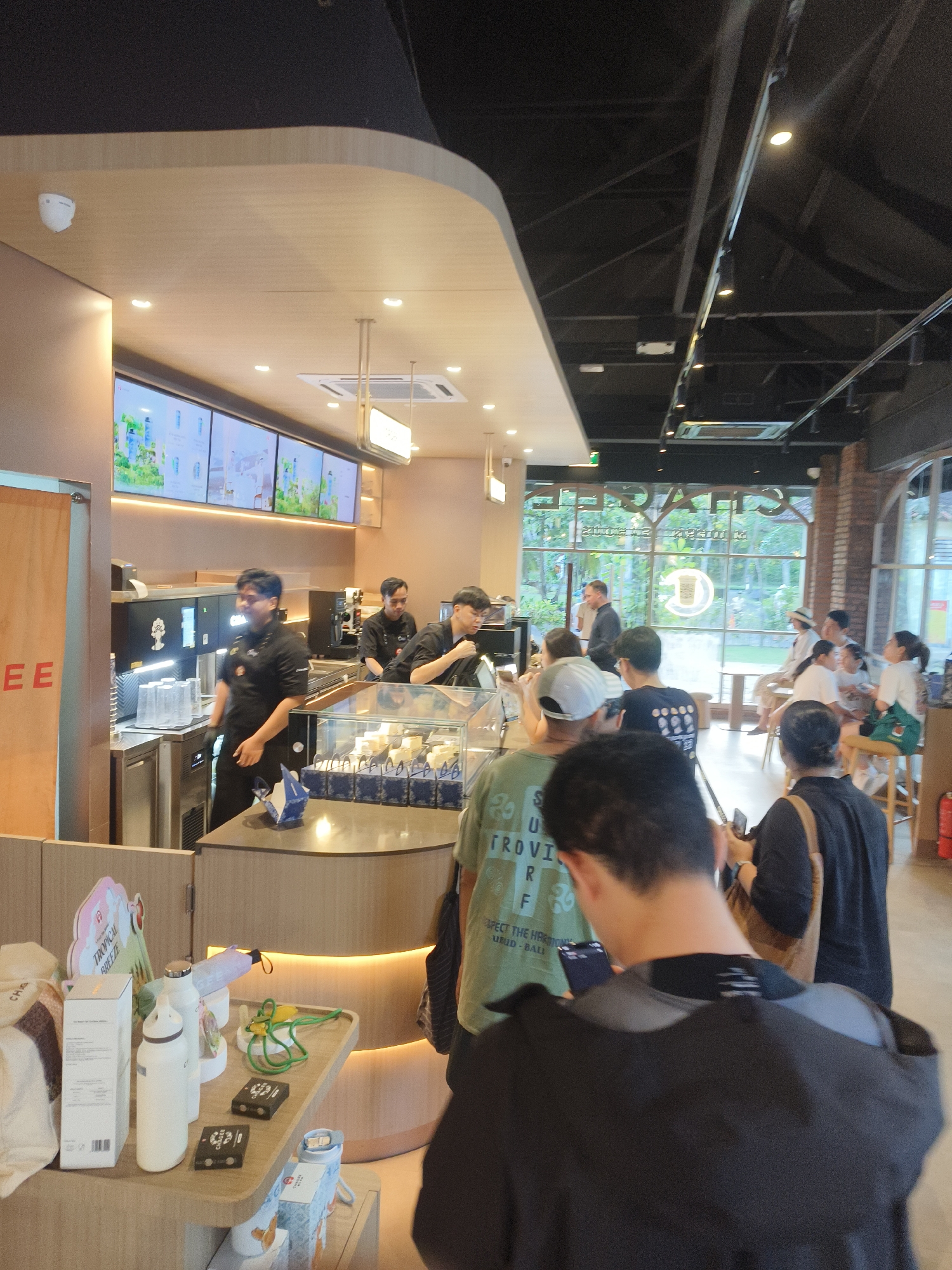 The image depicts the interior of a cafe, likely in Kuta Selatan, Indonesia, with a focus on the counter area and customers. The cafe has a modern design with light wood accents and a dark ceiling. Several employees in black uniforms are visible behind the counter, engaged in preparing drinks. A line of customers is queuing to order. On the counter, there are various items such as cups, drinks, and merchandise. In the background, through a large window, lush greenery and outdoor seating are visible, suggesting an outdoor ambiance or a view of a garden. The lighting is a mix of ambient ceiling lights and accent lighting, creating a warm and inviting atmosphere. A security camera is mounted on the ceiling. The overall scene conveys a busy and active cafe environment.