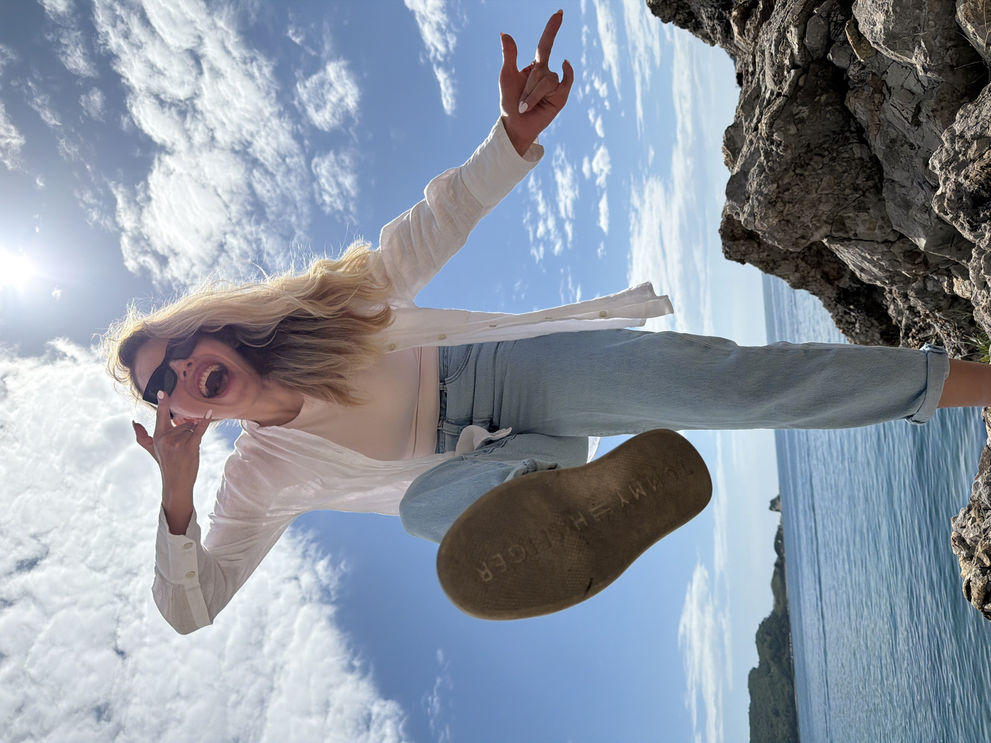 A blonde woman wearing a white blouse, jeans, and sunglasses is photographed from below. She is on a rocky cliff or overlook, with the Adriatic Sea and possibly Sveti Nikola Island visible in the background. The photo could have been taken in or near Budva, Montenegro, due to the coastal landscape. The woman is in motion, with one leg extended towards the camera, showing the sole of her Tommy Hilfiger shoe. She is making hand gestures, and her mouth is open in a shout or laugh. The sky is blue with scattered clouds, and the sun is bright. The overall impression is a playful and energetic moment captured on a sunny day.