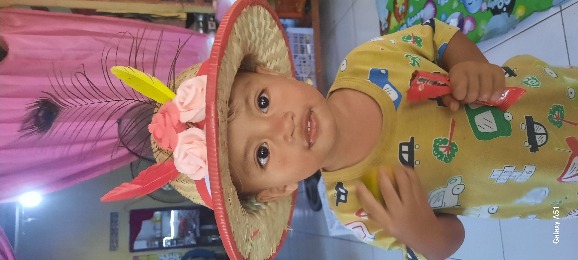 A young child wearing a straw hat decorated with fabric flowers and feathers is shown from the chest up. The hat has a red rim and straw-colored weaving. The child is wearing a yellow t-shirt with various cartoon vehicle prints. The child's right hand is holding a small red package, possibly candy. The background is out of focus, showing what appears to be an indoor setting with pink curtains, a light fixture, and some furniture. The image has a "Galaxy A51" watermark in the bottom left corner, indicating the device used for the photograph. The scene is consistent with a domestic setting within Ploso, Indonesia.