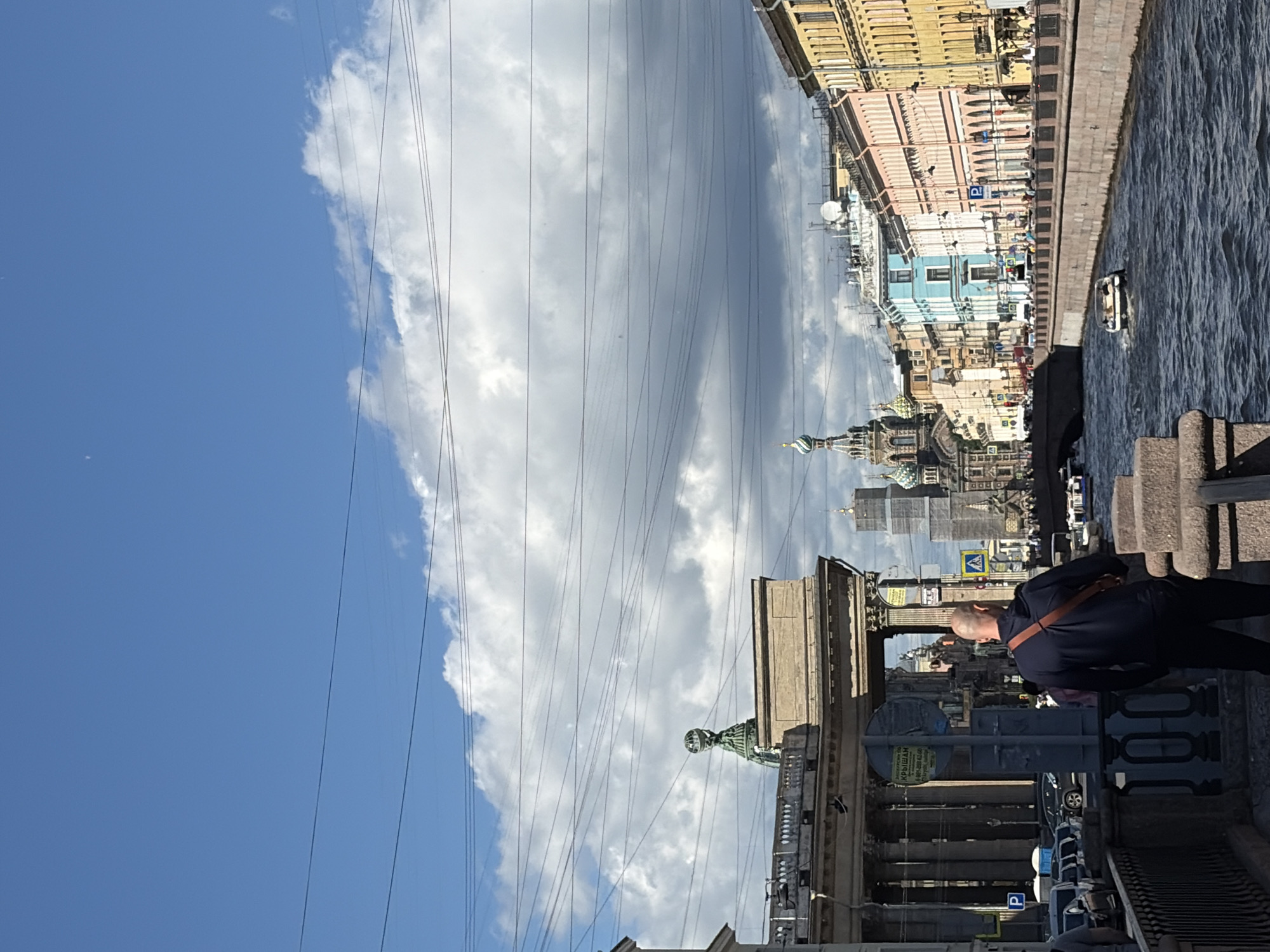The image depicts a street scene in Saint Petersburg, Russia, on a bright, partly cloudy day. The perspective is from a person standing on a bridge or embankment overlooking a canal or river lined with colorful buildings.
The foreground features a man with a shaved head, wearing a dark jacket and dark pants, with a light-colored strap across his shoulder. He is looking out at the scene, appearing contemplative. To his left, a stone balustrade runs along the edge of the waterway.
Beyond the man, the canal is filled with shimmering water, and a small white boat is visible. The buildings lining the opposite bank are tall and ornately decorated, painted in shades of yellow, pink, and blue. Some architectural details suggest historical significance, including domes and intricate facades.
A prominent feature of the image is the network of overhead wires crisscrossing the sky. The sky itself is a vibrant blue, punctuated by large, fluffy white clouds that cast shadows on the buildings below.
In the background, more of the city's architecture is visible, including church domes and modern buildings. Traffic signs, including one with a blue "P" indicating parking, are present along the street. Some Cyrillic text is visible on signs, though it is not fully legible. The overall impression is one of a bustling urban environment bathed in pleasant daylight.