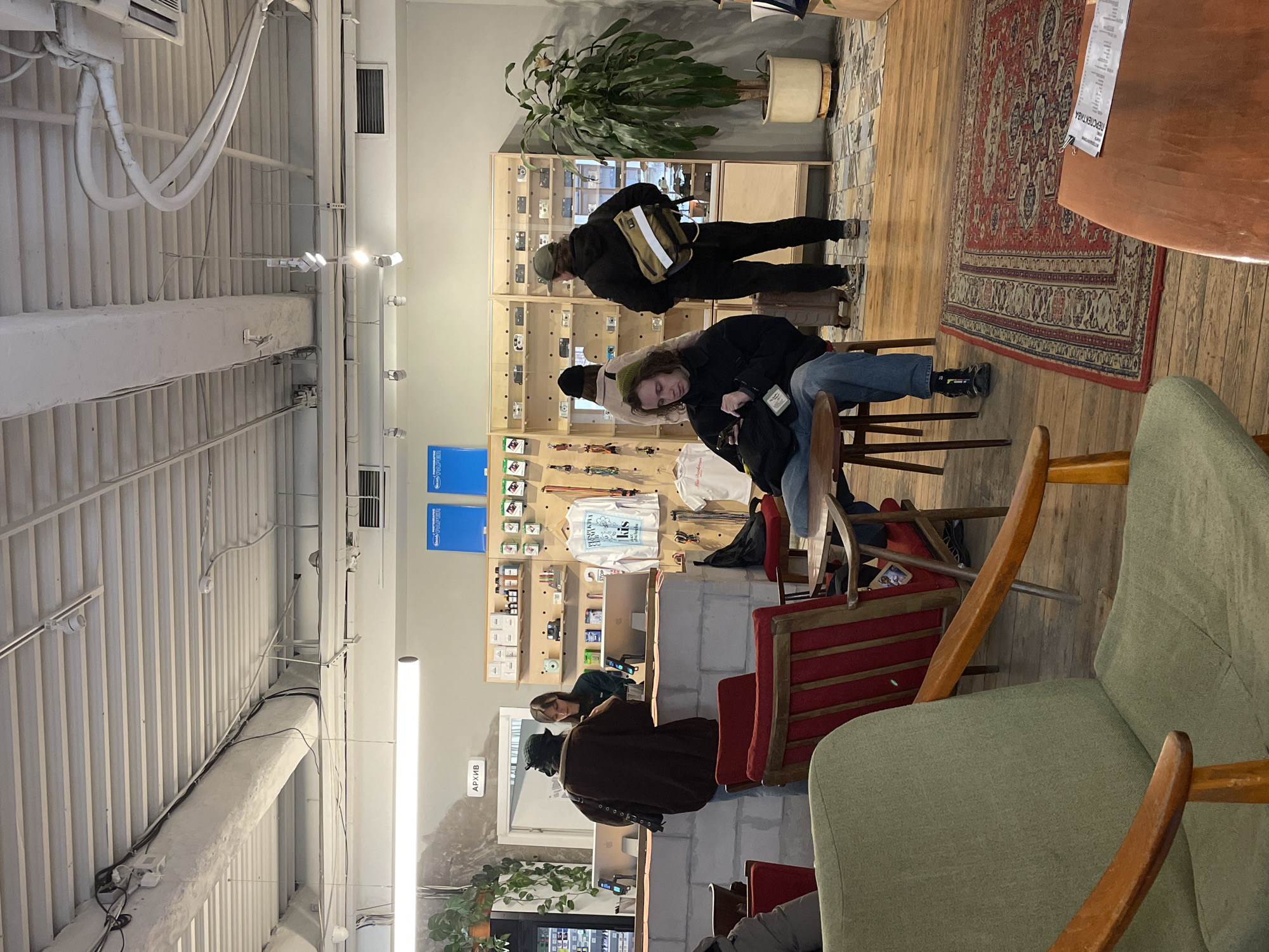 The image captures the interior of a retail space in Moscow, Russia. The shop, recognizable by signage in Russian, is populated with several individuals. Some customers are browsing a display of camera equipment, while others are interacting with staff at a counter. The decor features a mix of modern and vintage elements, including exposed ceiling fixtures, wooden flooring, and a vintage rug. Comfortable seating, such as upholstered chairs, is arranged in a seating area. The scene conveys a relaxed and engaged atmosphere within a niche retail environment.