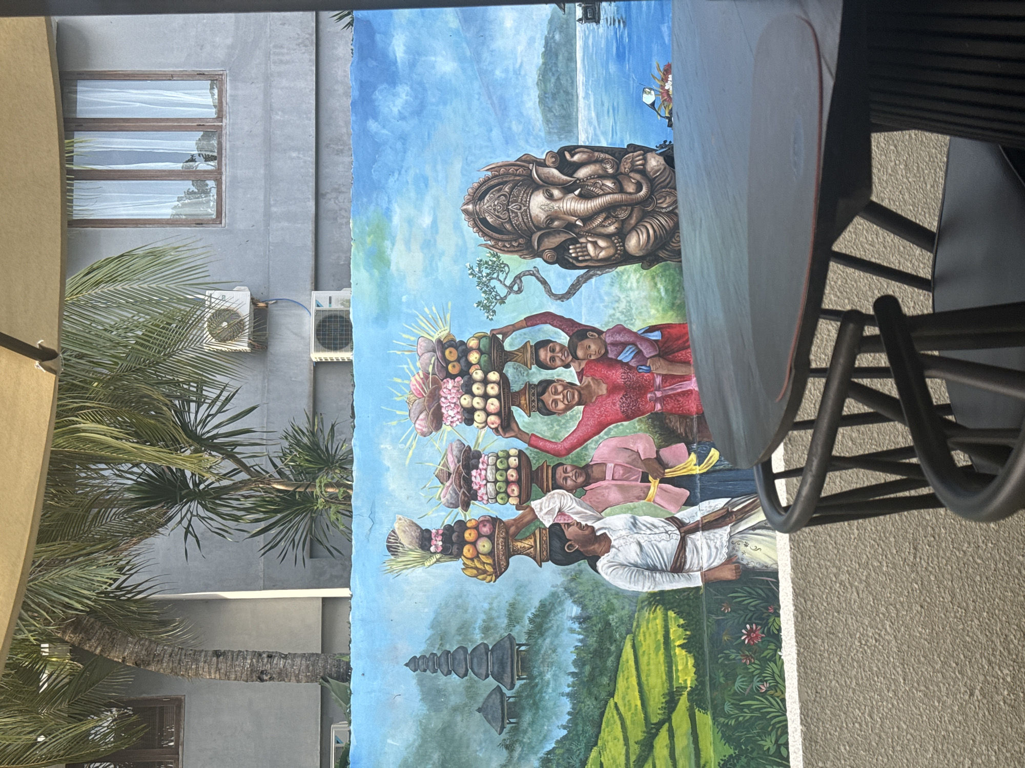 The image depicts an outdoor scene likely located in Kuta Selatan, Indonesia, with a mural painted on a wall. The mural features a depiction of Balinese life, including women carrying offerings on their heads, a Ganesha statue, and a landscape with rice paddies and a temple. In the foreground are a table and chairs, partially obscuring the mural. A palm tree is visible to the left of the mural, and a building facade is behind it, with windows and air conditioning units. The overall scene suggests a tropical, cultural setting.