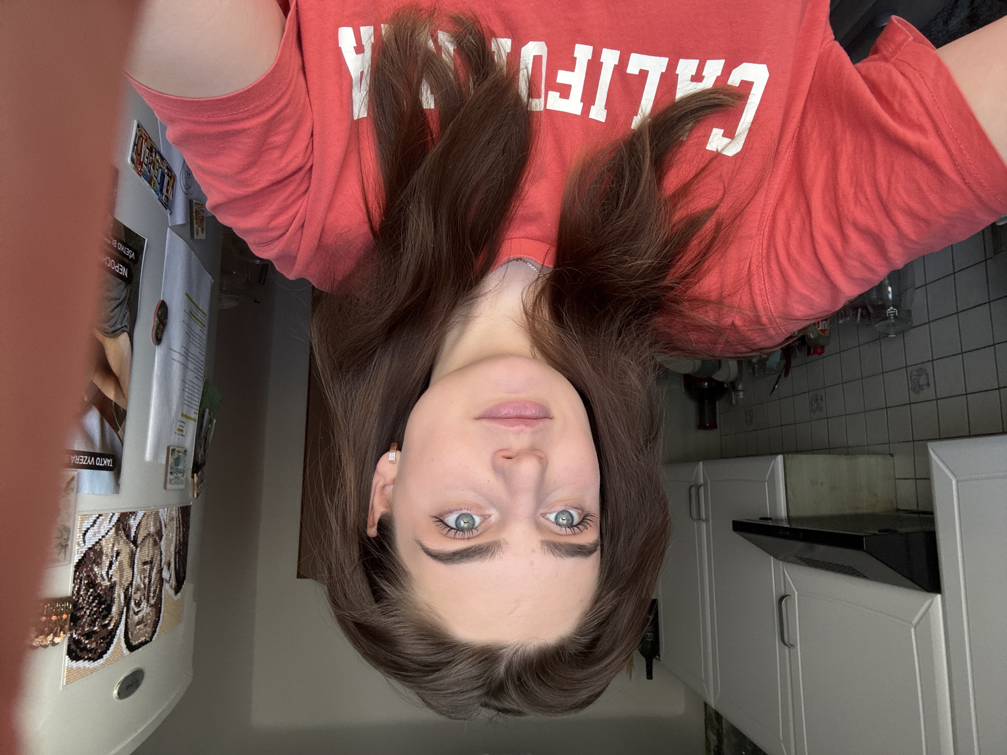 A selfie captures a young woman, inverted, looking directly at the camera with a neutral expression. Her long, dark brown hair frames her face and spreads across her shoulders. She has light blue-green eyes and is wearing small golden hoop earrings. Her eyelashes are prominent, suggesting mascara. She wears a reddish-orange t-shirt with large white uppercase letters that spell "CALIFORNIA" across the chest.
The background shows an indoor domestic setting, likely a kitchen. To the left (in the inverted image), a white refrigerator is visible, adorned with magnets and papers. Notable magnets include one resembling a car license plate with "ROZMARIN" printed on it, and another small, dark, possibly shoe-shaped object. Visible text on papers attached to the fridge includes Slovak phrases like "TAKTO VYŽERA" (meaning "THIS IS WHAT IT LOOKS LIKE") and "NEPOCH" (likely part of "nepochopitelné," meaning "unbelievable" or "impossible"), along with "VŠETKO BL".
To the right of the woman, white kitchen cabinets with silver handles are arranged along a wall, with a black range hood mounted above what appears to be a cooktop area. Light-colored tiles are visible above the cabinets. A dark bottle, possibly wine, is seen near the bottom right. The lighting is artificial and bright, suggesting an indoor environment. The presence of Slovak text on the refrigerator items is a notable cultural detail, fitting with the location context of Hainburg an der Donau, Austria, which is close to the Slovak border. The woman's posture and the inverted image suggest she might have been lying down while taking the photo.