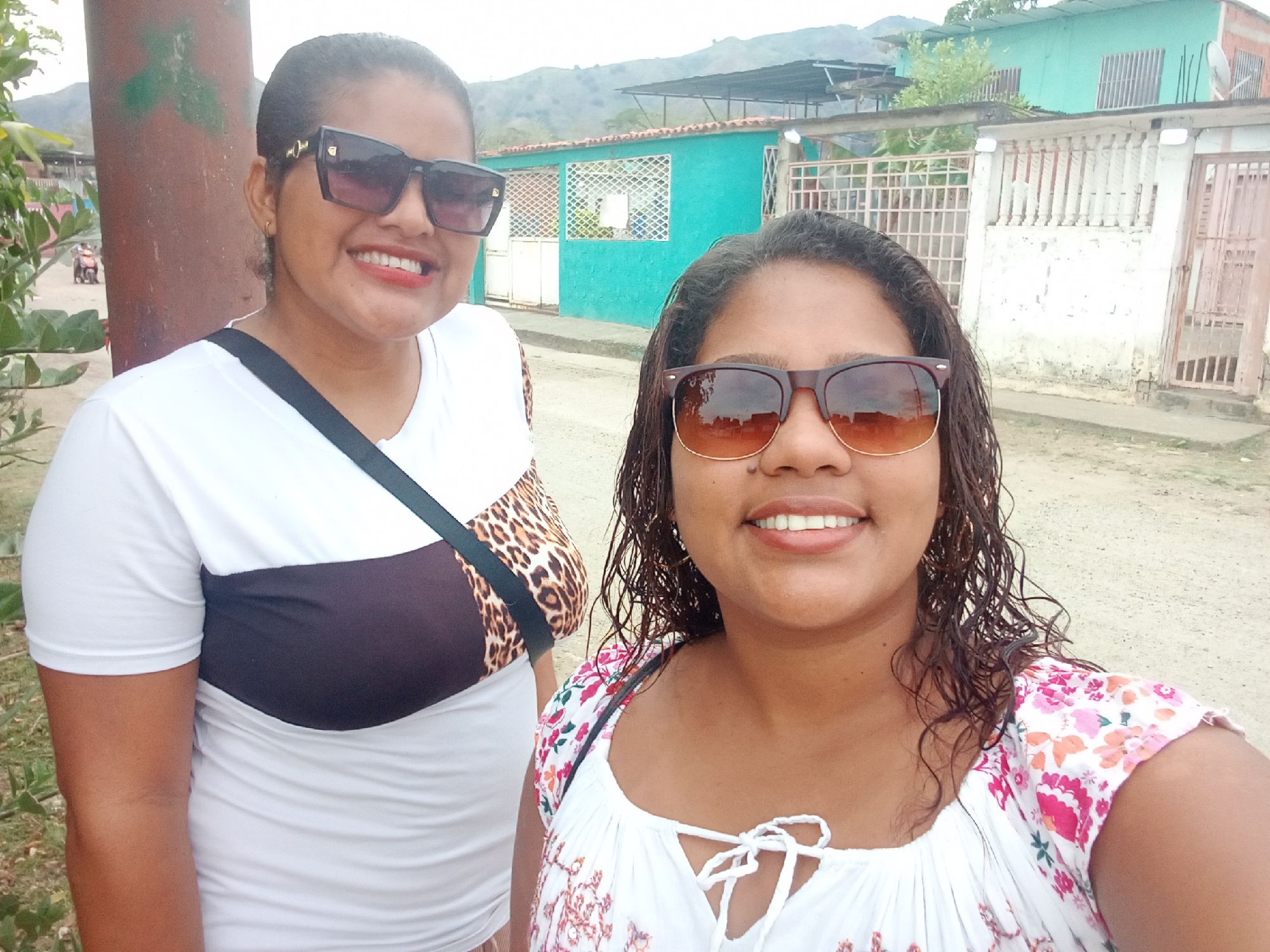 Two women are posing for a photograph on a street in Parroquia Ocumare, Venezuela. The woman on the left wears a white t-shirt with brown and leopard print panels, dark sunglasses, and a black strap across her chest. The woman on the right wears a white floral print top and brown sunglasses. In the background is a turquoise building and a white fence. A utility pole stands to the left of the women. The street is unpaved. Mountains are visible in the distance.