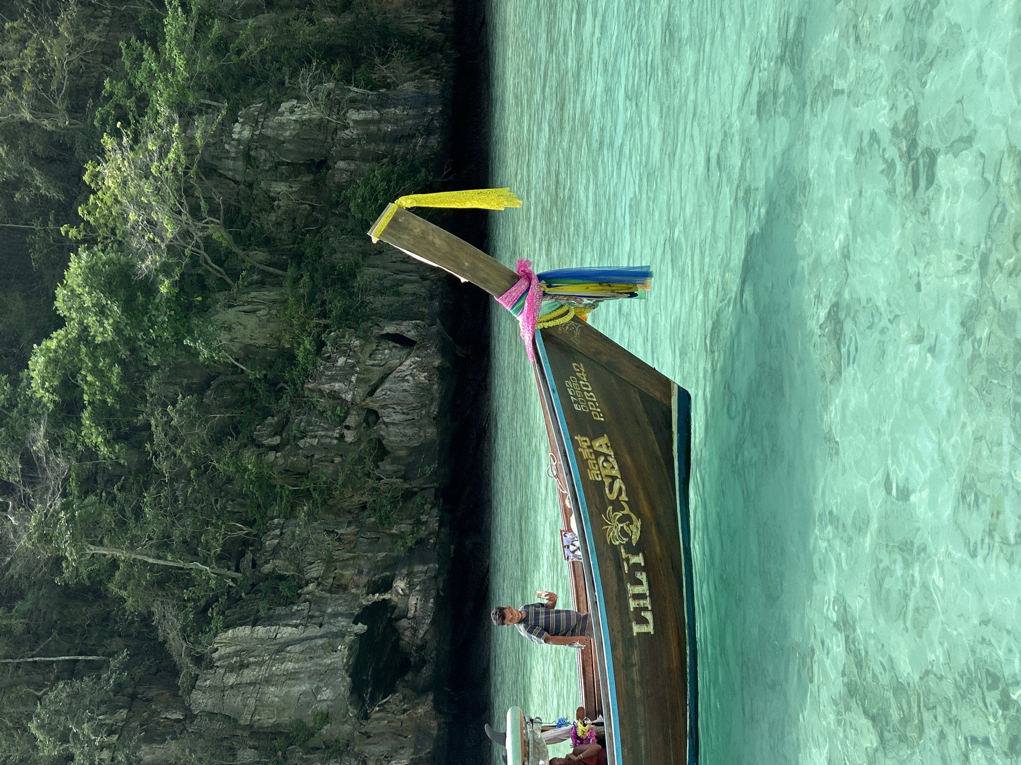A wooden longtail boat, named "LILY SEA," floats on the clear turquoise waters of Ao Nang, Thailand, near a lush green cliff face. A young man in a striped shirt stands in the boat, holding a small white object. The boat is decorated with colorful ribbons and has a Thai script on its side. The water is shallow enough to see the seabed, and the sunlight creates ripples on the surface. The scene conveys a sense of tropical tranquility.