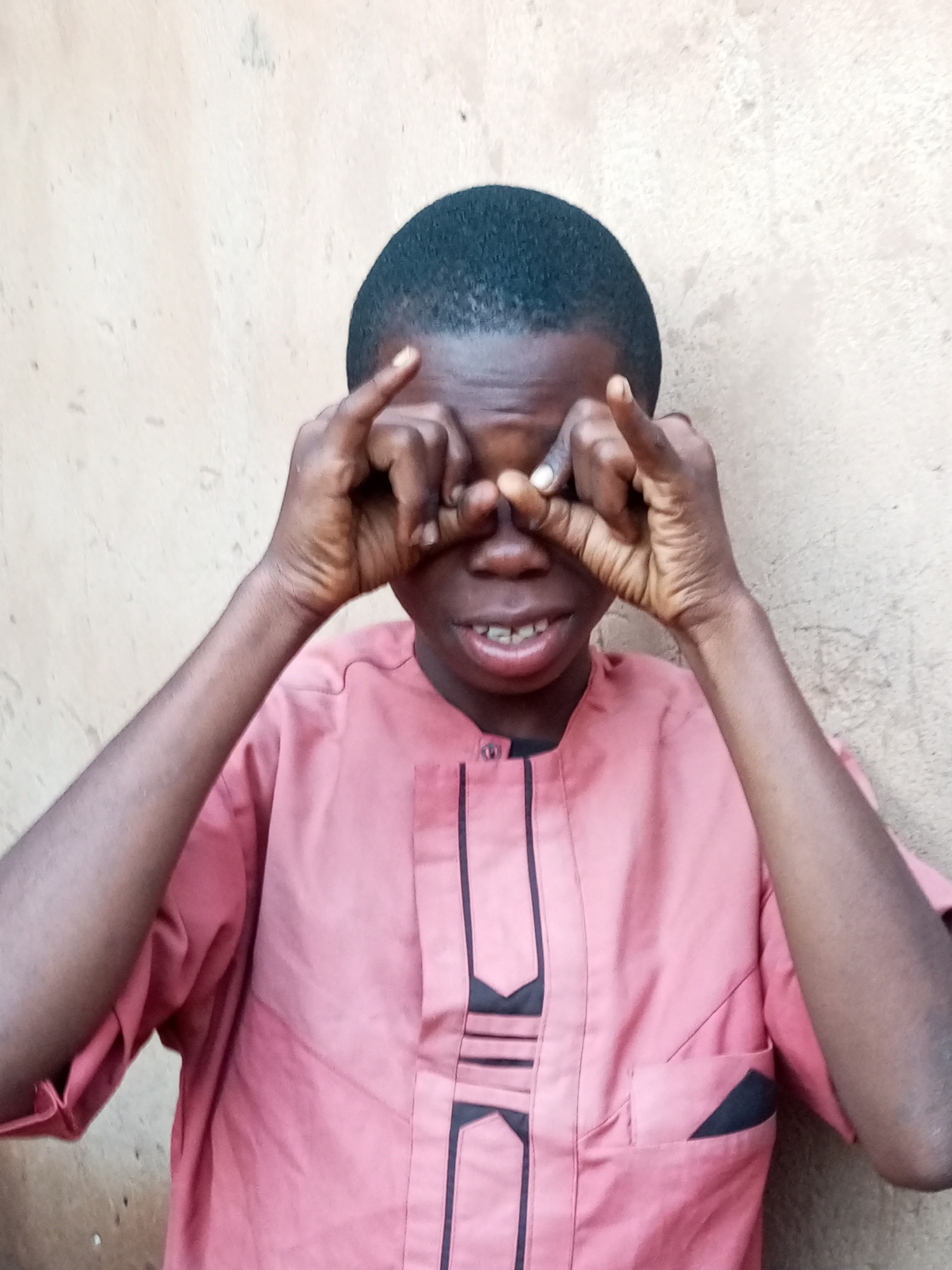 A young Black boy, appearing to be around ten years old, is shown in a close-up, front-facing portrait against a textured, beige wall. He has short, dark hair and is wearing a pink, collared shirt with dark trim and detailing. The boy is looking directly at the camera with his eyes covered by his hands, creating a playful or curious expression. His fingers are interlaced over his eyes, with his thumbs positioned on his cheeks and index fingers extended upwards. The background suggests an outdoor urban setting, likely in Tempure, Nigeria. The lighting is even, indicating it could be daytime.