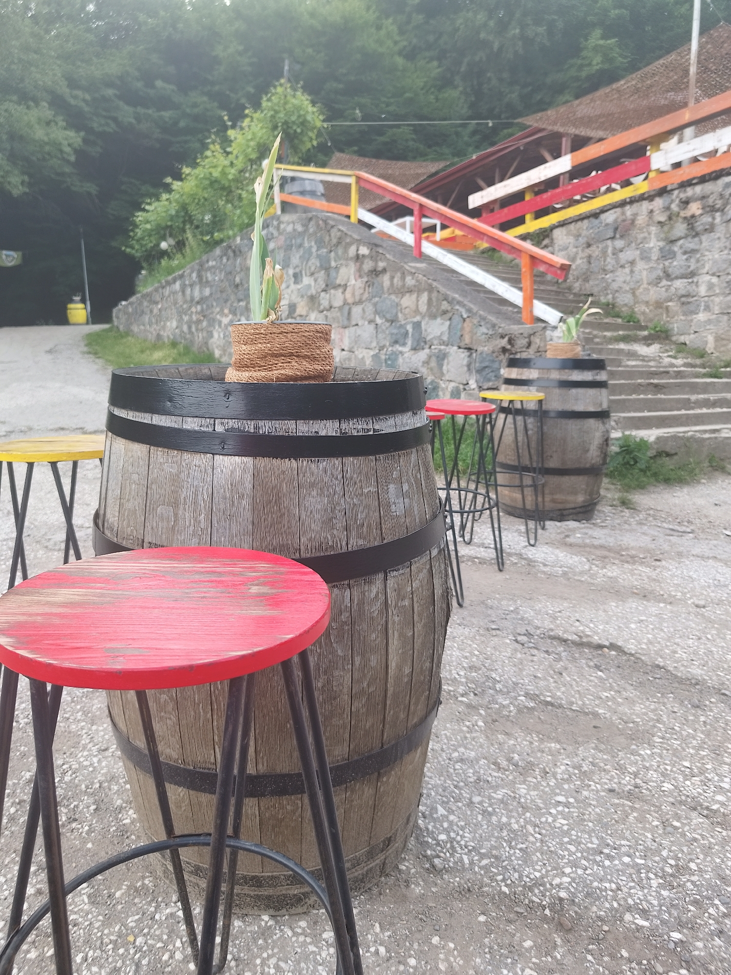The image shows a wooden barrel with a plant in a woven pot on top, placed in an outdoor setting. In front of the barrel is a red stool. To the left and right of the barrel are other stools, one yellow and two more visible further back. The barrel and stools are set on a gravelly surface. Behind them, a stone wall with steps leads up to a building with a red roof and a colorful railing. The railing has alternating sections of white, yellow, red, and orange. The background features a dense forest. The overall scene suggests an outdoor seating area, possibly at a restaurant or bar, with a rustic ambiance. Based on the context provided by the user, this scene is located in the city of Sofia, Bulgaria.