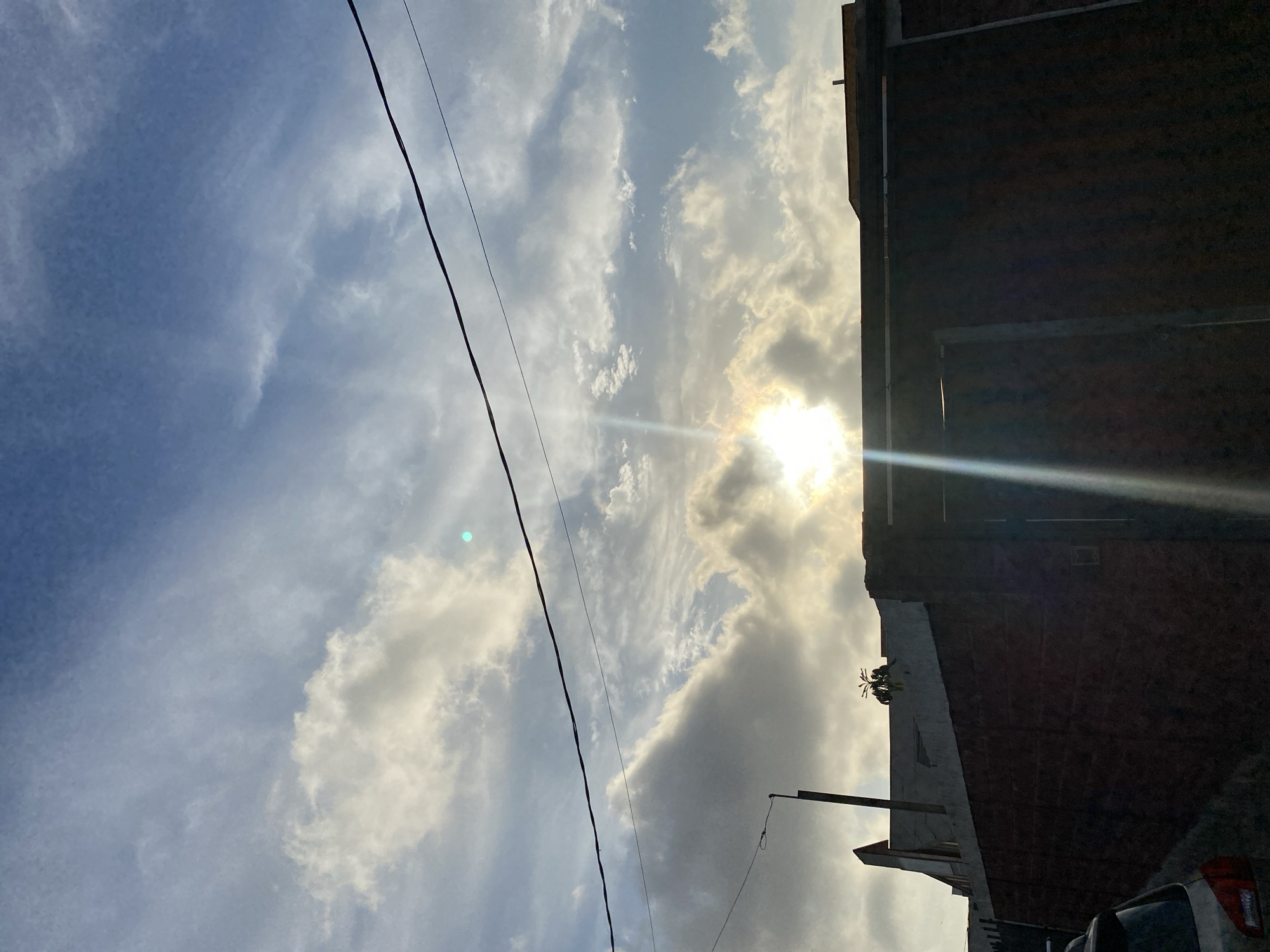 The image, taken in Arica, Chile, is a street-level upward view. The sky is mostly blue with some white clouds, through which the sun is shining. The clouds range from thin and scattered to denser formations. In the foreground, a dark electrical wire runs diagonally across the scene. On the right, the side of a brick building is visible, blocking some of the sky. At the base of the building, some smaller buildings are visible. A parked car can be seen at the lower right corner. A street lamp post is also present.