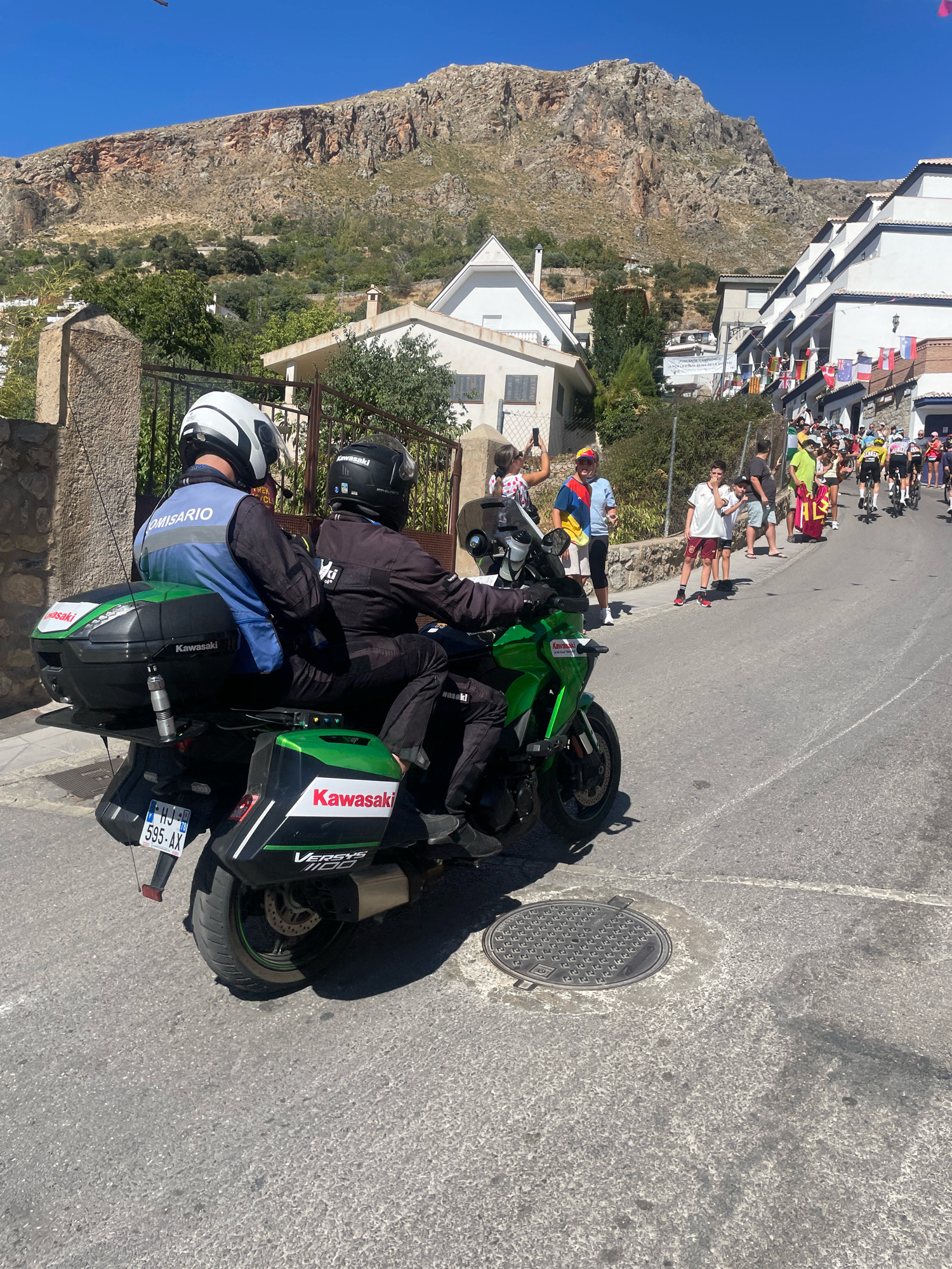 In Güéjar Sierra, Spain, on a sunny day, two individuals in motorcycle gear, identified as "COMISARIO" on the vest of the front rider, are seated on a green Kawasaki motorcycle. The motorcycle is parked on a paved street, with its rear storage compartments visible, one bearing "Kawasaki" branding and "VERSYS 1100". The license plate "HJ 595-AX" is affixed to the rear. In the background, a hillside community with white buildings is visible, adorned with numerous flags. A crowd of spectators, some appearing to be cyclists in racing attire, lines the street. The mountainous terrain of Güéjar Sierra forms the backdrop under a clear blue sky.