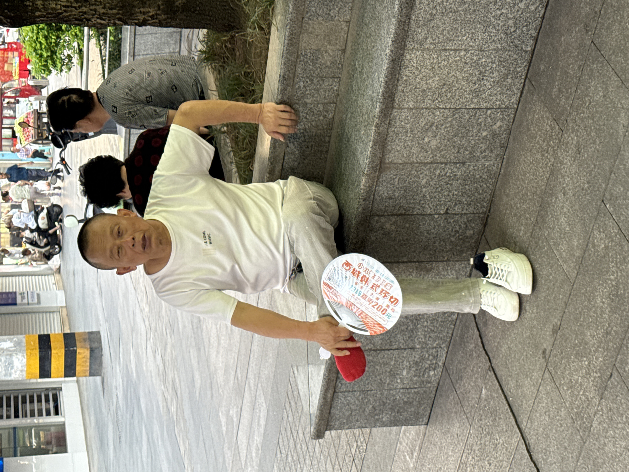 This media file captures an outdoor scene in an urban environment, likely a bustling street or walkway in Jiangjin, China, during daylight hours. The main subject is an older man, bald or with very short hair, wearing a white t-shirt with "E COML MUSIC" printed on it, and light-colored pants. He is sitting on a low grey speckled stone wall or curb, looking towards the camera with a slight smile.
In his right hand, he holds a round white and red promotional fan. Visible text on the fan includes "西城韩式环切" (Xicheng Korean Style Circumcision), alongside "安全 | 美观" (Safe | Beautiful) and an offer "扫码直降200元" (Scan code for a direct discount of 200 yuan). A red object, possibly a small bag or folded item, rests on his leg. He is wearing white sneakers.
Behind him, two other individuals are partially visible: one person in a grey patterned shirt and another in a dark red patterned shirt. In the background, a busy street scene unfolds with several pedestrians and a red fruit stall. A black and yellow striped barrier is also visible on the left side of the frame. The presence of trees and greenery suggests a public area. The bright, even lighting indicates clear, possibly warm, weather.