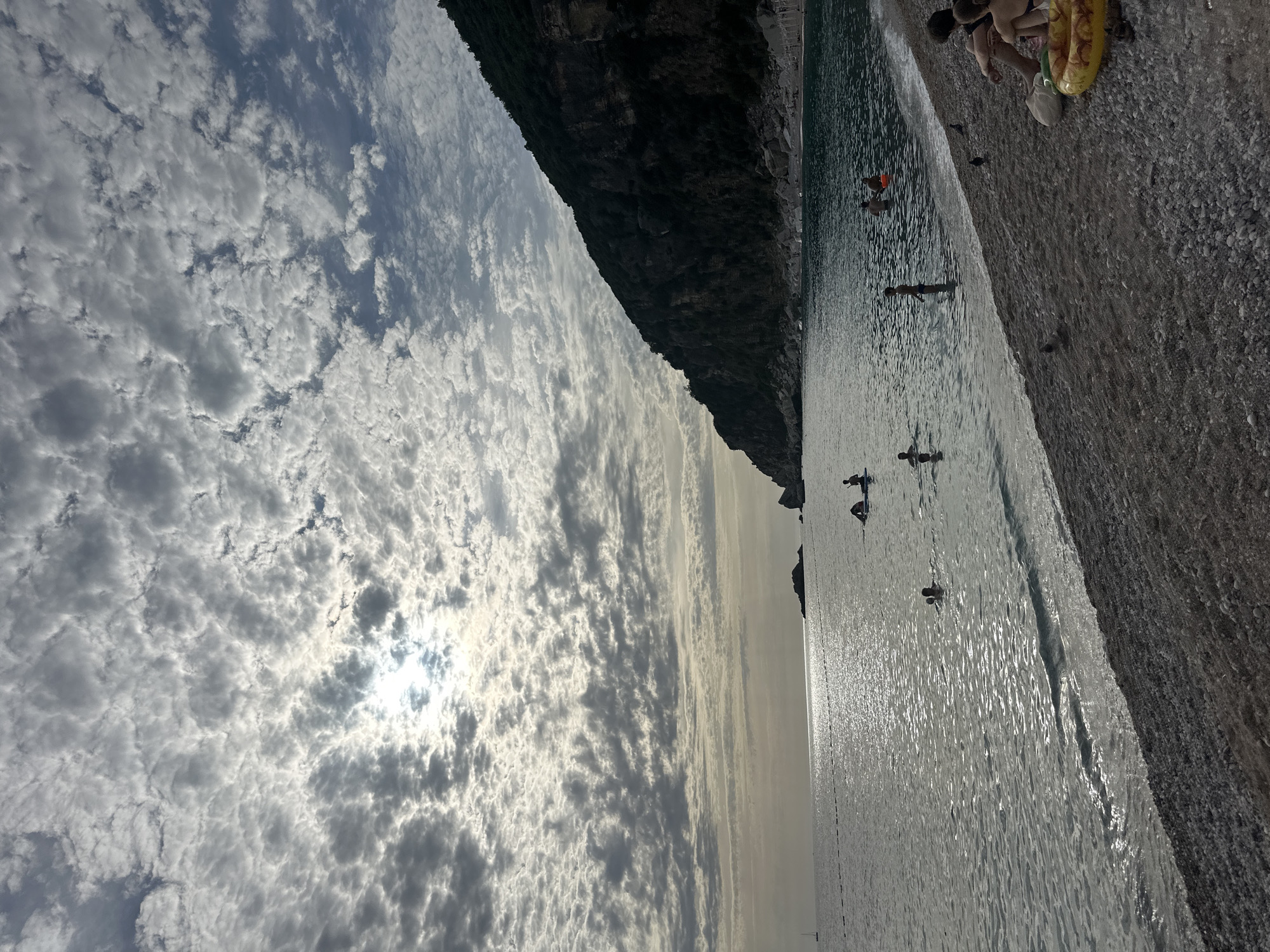The image depicts a crowded pebble beach and the sea at Mirište, Montenegro, under a heavily clouded sky with the sun breaking through. The sky is largely covered by numerous small, dense grey clouds, with a bright patch indicating the sun's position slightly left of the image center, creating a strong backlight. This illumination causes a shimmering path of reflected light on the water surface, extending from the horizon towards the viewer.
To the right, a large, dark, rocky cliff covered with sparse vegetation slopes steeply into the sea, appearing silhouetted against the brighter sky and water. A line of white buoys or floats stretches horizontally across the mid-ground of the water, marking an unseen boundary. In the far distance on the left, a small sailboat is visible.
Multiple individuals are present in the scene. In the water, at least seven people are visible: two are on paddleboards or similar small craft near the right shore, one is wading further out to the left, and several others are swimming or standing in the water closer to the beach. In the immediate foreground on the pebble beach, two individuals are seated, partially silhouetted. A yellow inflatable swimming ring lies next to them. Three pigeons are visible on the pebbles near the seated figures, and another is closer to the water's edge. The overall lighting suggests either late afternoon or early morning, with elements of the landscape and figures appearing silhouetted.
