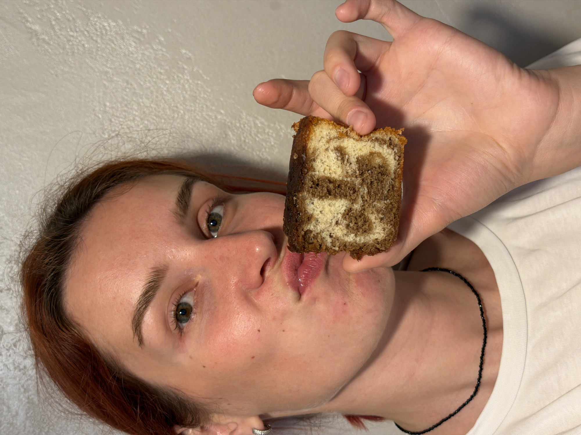A woman with reddish-brown hair is holding a square piece of marbled cake in front of her face. She is looking at the camera. The background is a plain white wall. She is wearing a white shirt and a black beaded necklace. There is no information to suggest the location is Naberezhnye Chelny, Russia.