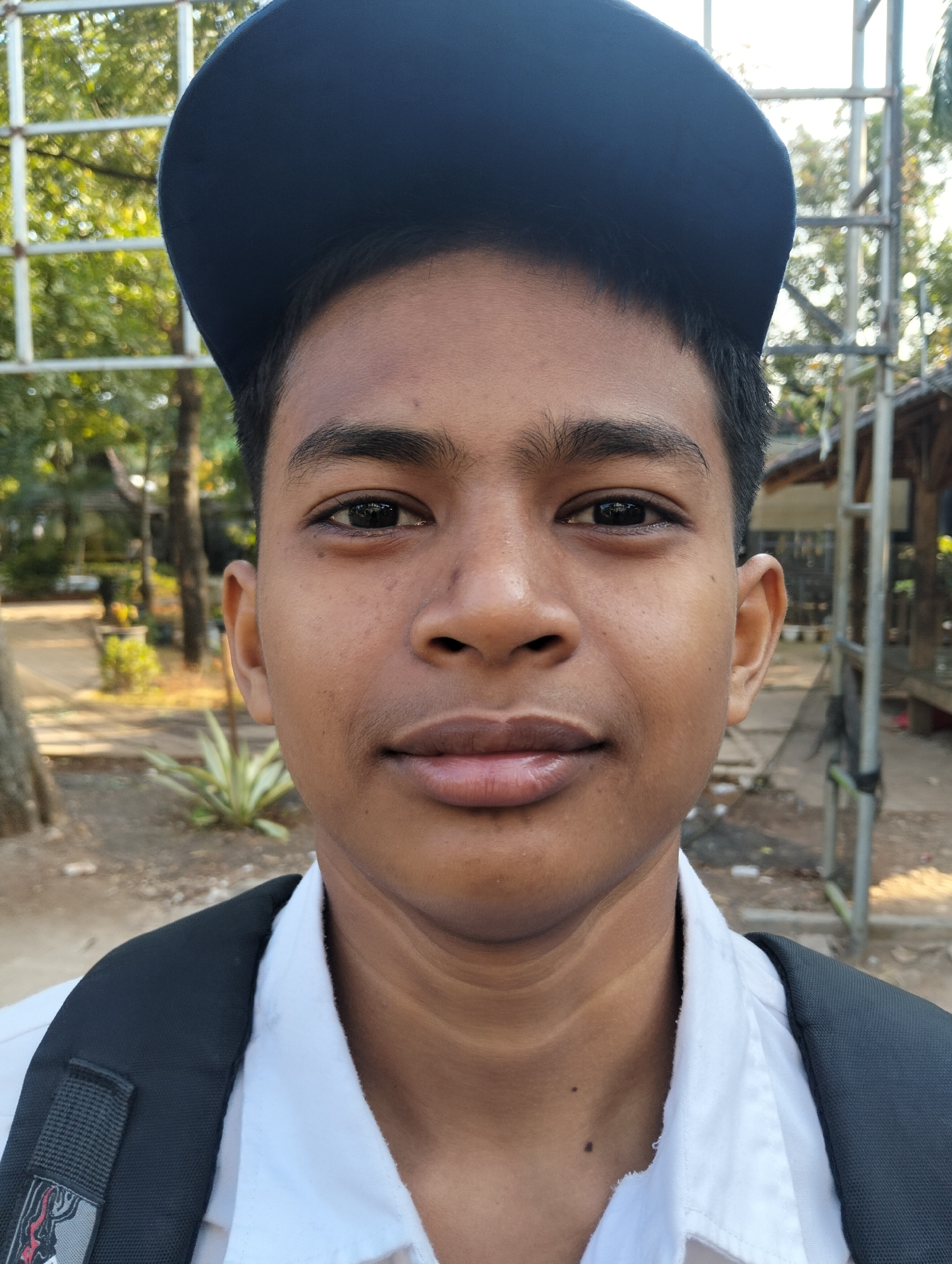 The image is a close-up, front-facing portrait of a young male, likely a teenager, wearing a dark blue baseball cap and a white collared shirt with a dark backpack strap visible over his shoulder. He has dark hair and a smooth complexion. The background is blurred, showing trees and some outdoor structures, suggesting an open-air setting. Based on the provided context, this scene is located in Paciran, Indonesia. The lighting indicates it is daytime.