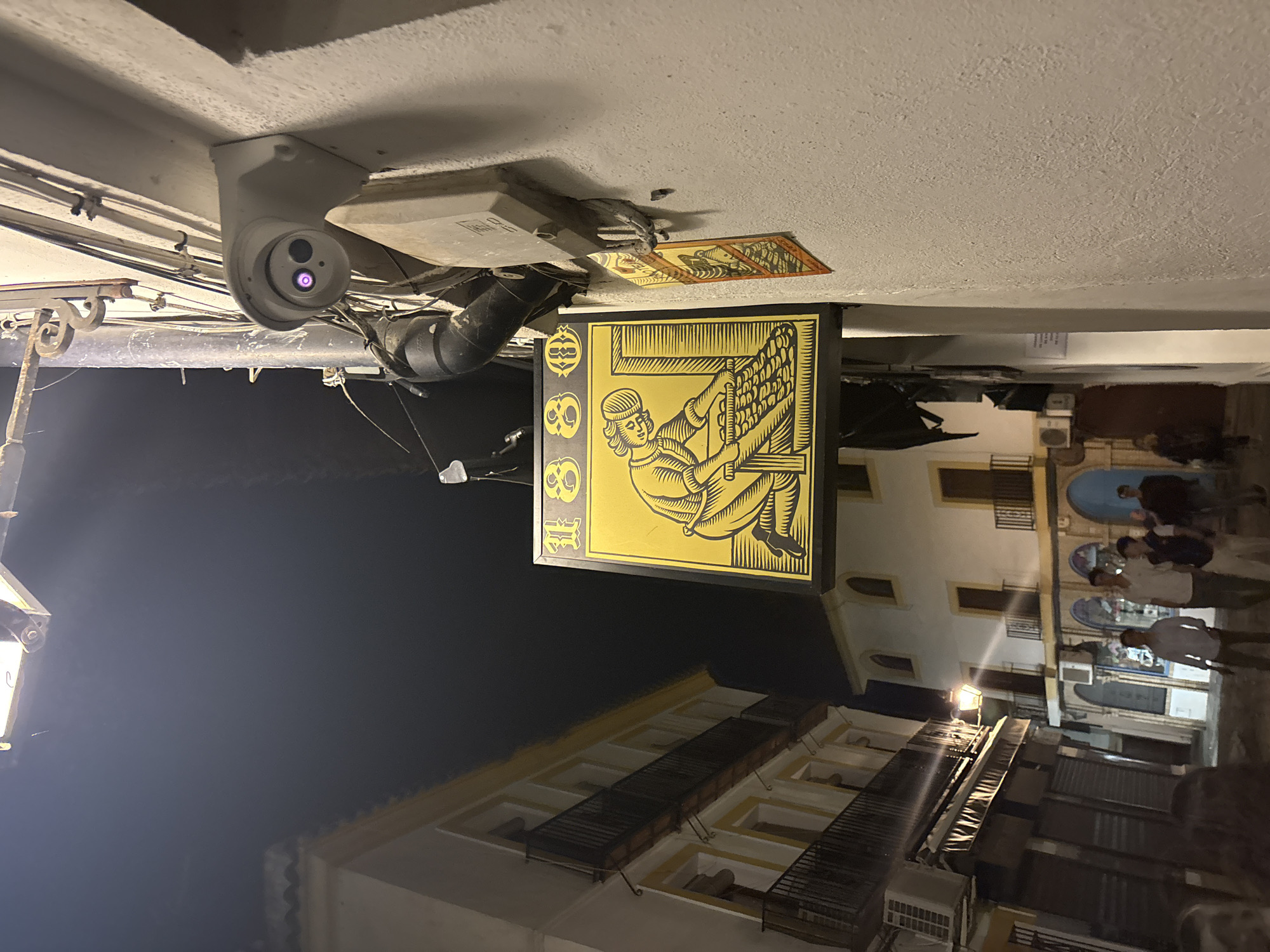This is a nighttime urban scene in Córdoba, Spain. The image is angled upwards, focusing on the architecture and street life.
A security camera is mounted on the ceiling of a building. The building also features a sign with a historical motif. The sign displays a yellow illustration of a figure using a printing press, set against a black background.
The street below is lit by streetlights. People are walking on the street, appearing as blurred figures, indicating movement. The buildings lining the street are multi-story, with visible windows and balconies. The architectural style suggests a historic urban area.