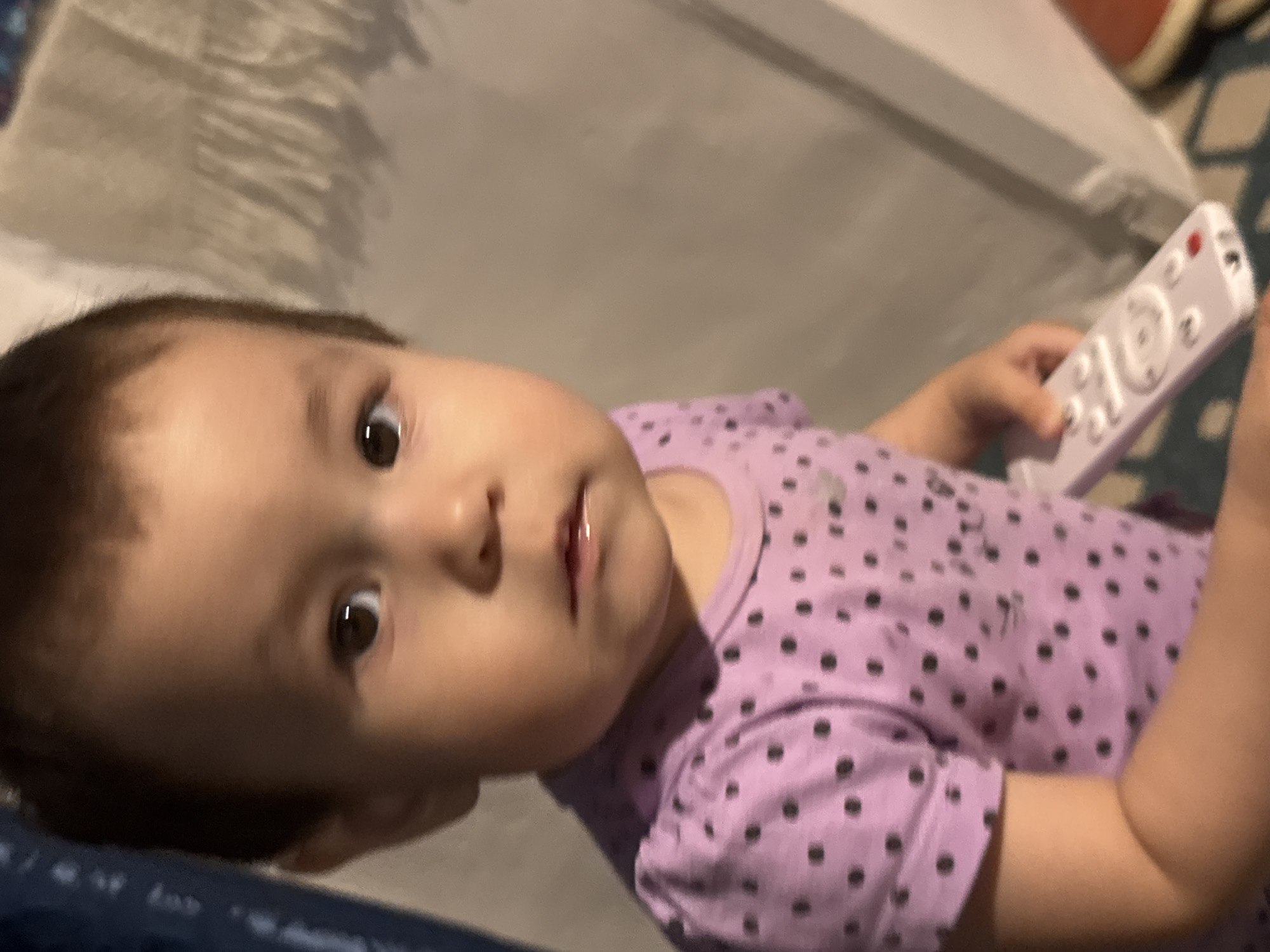 A close-up, eye-level shot shows a toddler, possibly of South Asian or mixed ethnicity, with dark hair, brown eyes, and fair skin. The child wears a purple t-shirt with black polka dots and is holding a white remote control. The child's mouth is slightly open, with a visible wetness, and their expression is neutral. The background is out of focus, but appears to be an indoor setting with a wall and a patterned rug. The overall lighting is soft and warm, with a slight shadow cast on the child's face. The image was taken in Chardara, Uzbekistan.