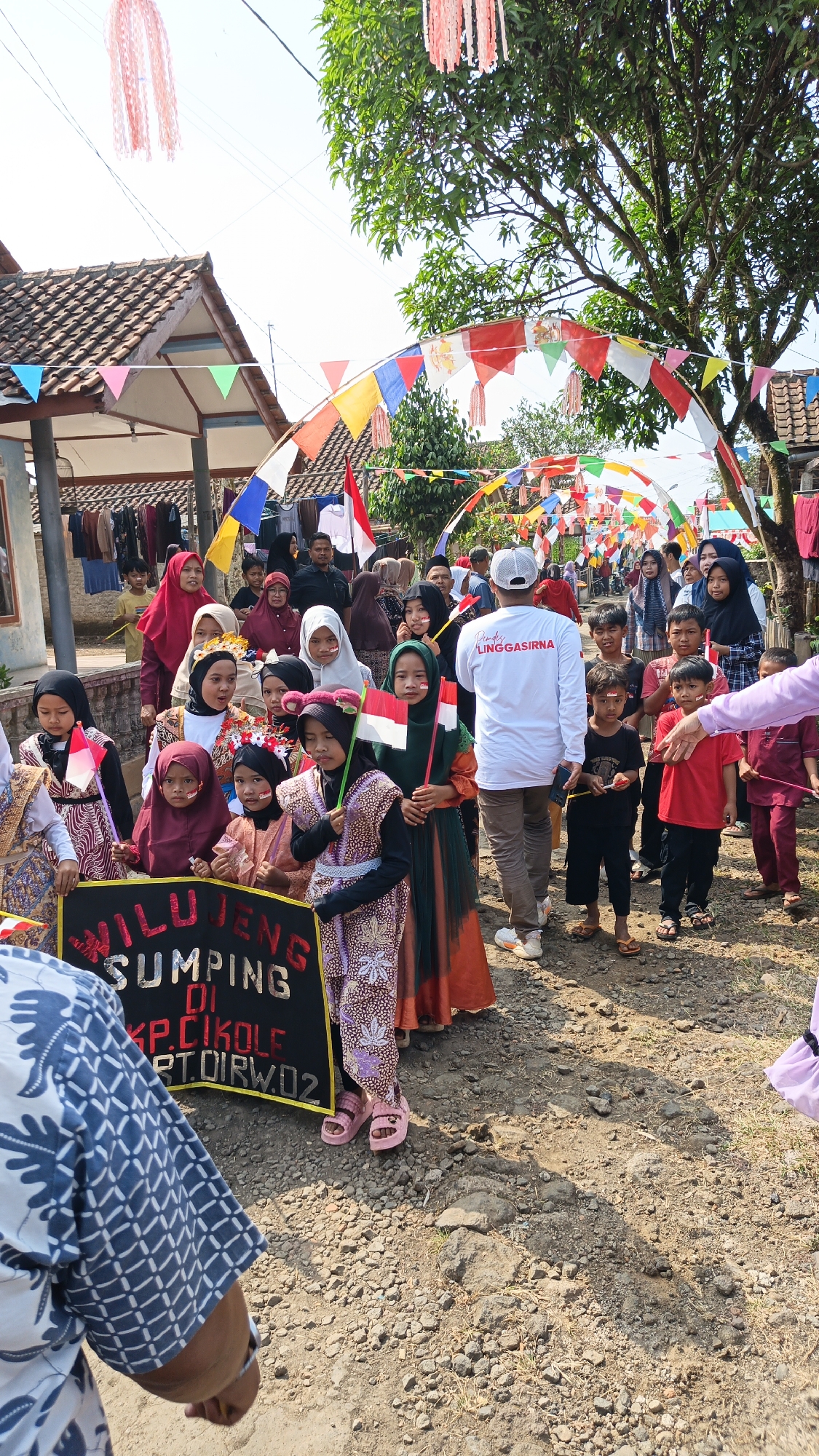The image depicts a street scene in Linggasirna, Indonesia, possibly during a local event or parade. A group of children, mostly girls dressed in traditional Indonesian attire and some holding small Indonesian flags, are gathered together. In the foreground, a black sign with yellow lettering is visible, partially obscured by a person wearing a blue and white patterned shirt. The sign appears to have text related to "Linggasirna" and "Kole".
Above the street, colorful festive decorations, including triangular flags and streamers, are strung across the area, forming an archway. A few adult men and women are also present in the background, observing or participating in the event. The environment appears to be a village setting, with houses and trees lining the dirt road. The overall atmosphere suggests a community gathering.