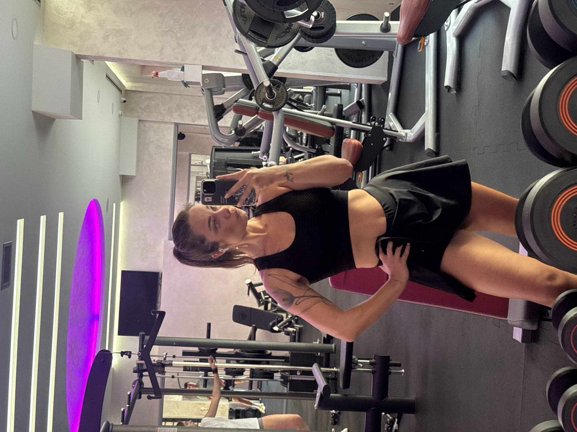 A woman with her hair in a ponytail, wearing a black sports bra and a black skirt, takes a selfie in a gym. She is holding a black smartphone in her right hand. Her left hand is on her hip. She has tattoos on her left arm and shoulder. The gym has weightlifting equipment, including dumbbells and weight racks. The floor is black rubber matting. In the background, another person is visible, also in gym attire. A television screen is mounted on the wall. Overhead, a purple light fixture illuminates part of the ceiling. The image was taken in Budva, Montenegro.