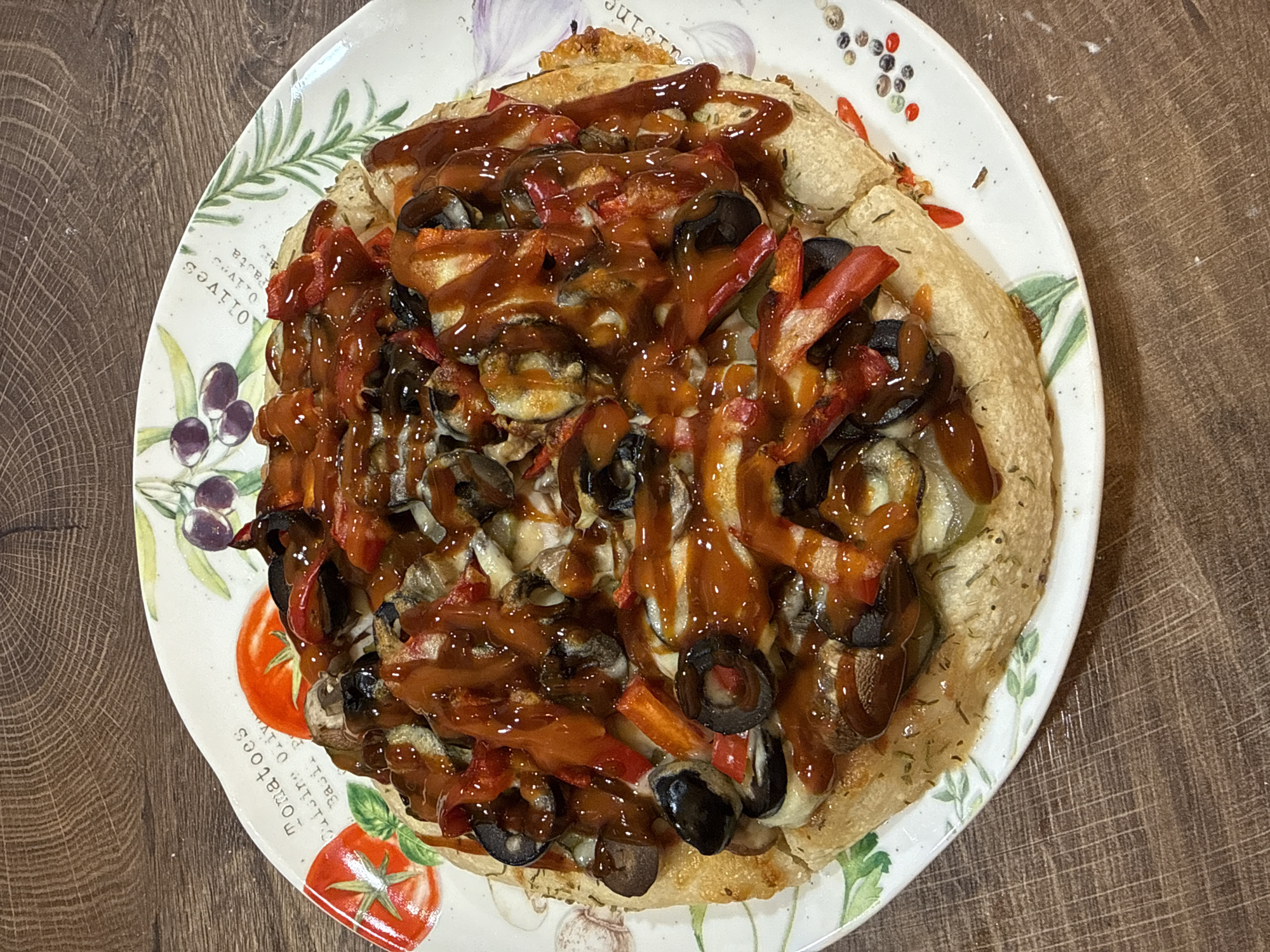 A top-down shot captures a round, savory dish, likely a pizza or flatbread, served on a decorative white ceramic plate set against a dark wooden table. The dish features a light golden-brown, herb-dusted crust with varied toppings. Visible ingredients include generous amounts of sliced black olives, strips of red bell pepper, and what appear to be mushrooms, all covered in melted cheese. A thick, glossy, reddish-brown sauce is liberally drizzled over the entire surface, creating a distinct visual pattern.
The ceramic plate's rim is adorned with colorful illustrations of culinary elements. On the left side, green olive branches with purple olives are depicted, accompanied by the visible text "OLIVES" and a partial "PASTA". Towards the bottom-left, red tomatoes with green leaves are illustrated, alongside the words "Tomatoes" and "basil". The wooden background exhibits a clear grain pattern. The scene is brightly lit, suggesting an indoor setting, and there are no people present.
