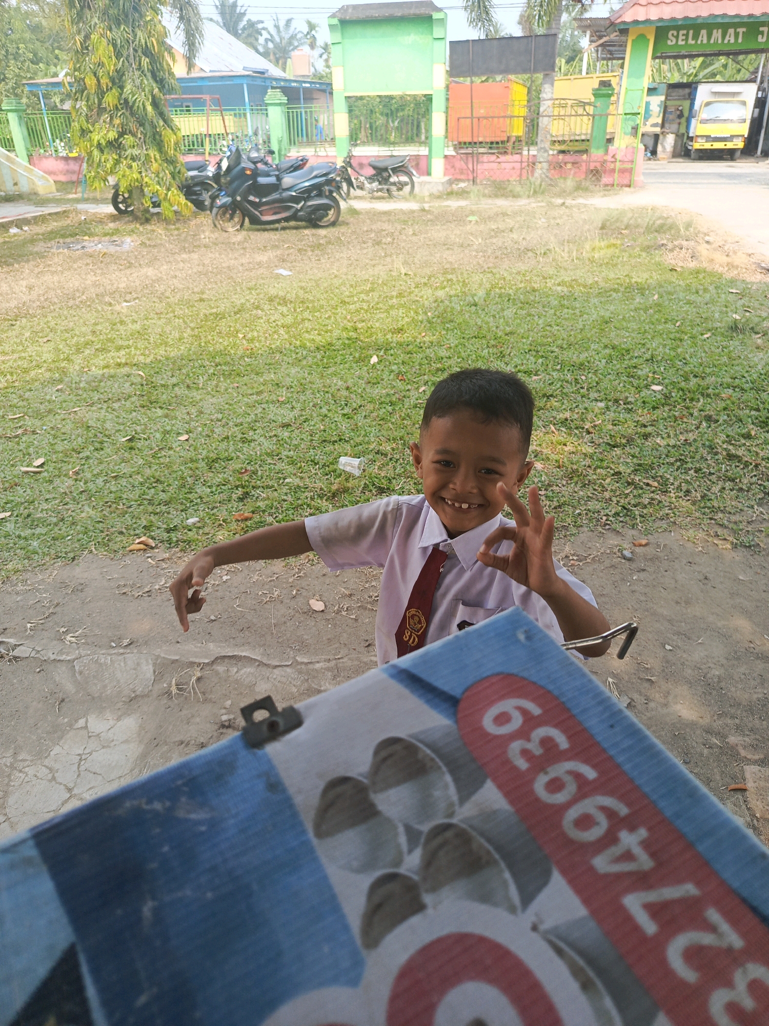 A young Indonesian boy, smiling and wearing a white school uniform with a red tie, is sitting outdoors. He is making an "okay" hand gesture with his right hand. In front of him, a blue and white patterned object with red lettering and numbers is partially visible. The background shows a grassy area, dirt ground, a tree, and a building with green and orange accents. Motorcycles are parked in the middle ground, and a yellow truck is visible in the distance, suggesting the location is near a street or possibly a checkpoint in Karya Dadi, Indonesia. The lighting indicates it is daytime.