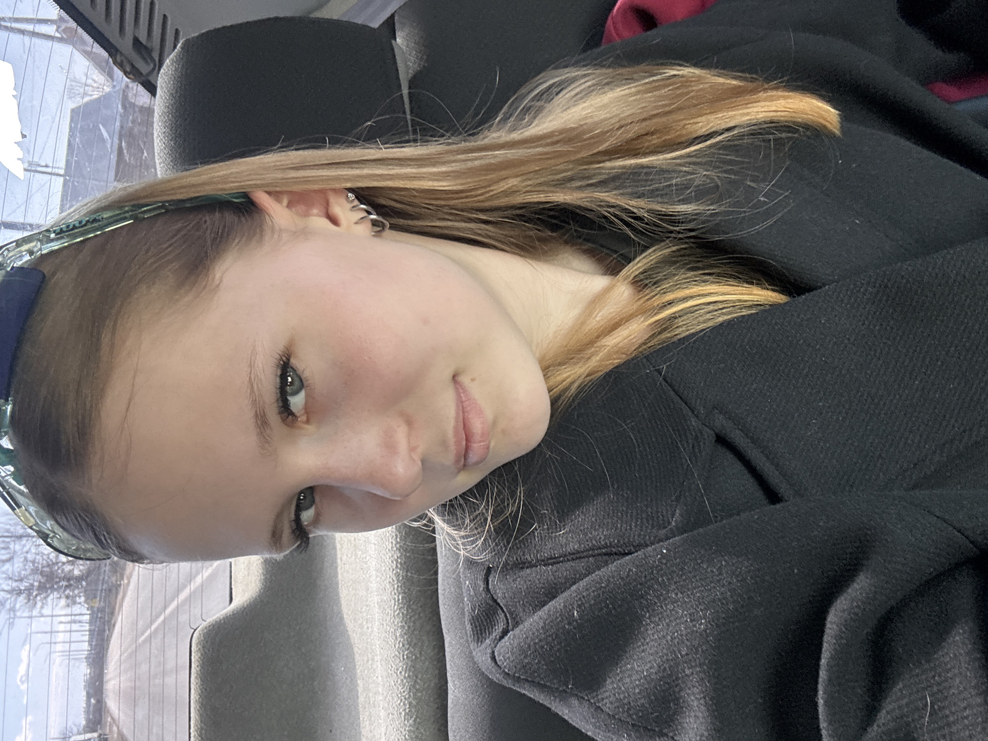 A fair-skinned young woman with light brown and blonde hair, wearing a black coat, is seated in what appears to be the backseat of a car. She has a headband and multiple earrings in her left ear. The background is blurred, showing a glimpse of a cityscape outside the car window. The location is potentially in the city of Кварса, Russia, based on the provided information. The specific actions and interactions are not directly observable from the image.