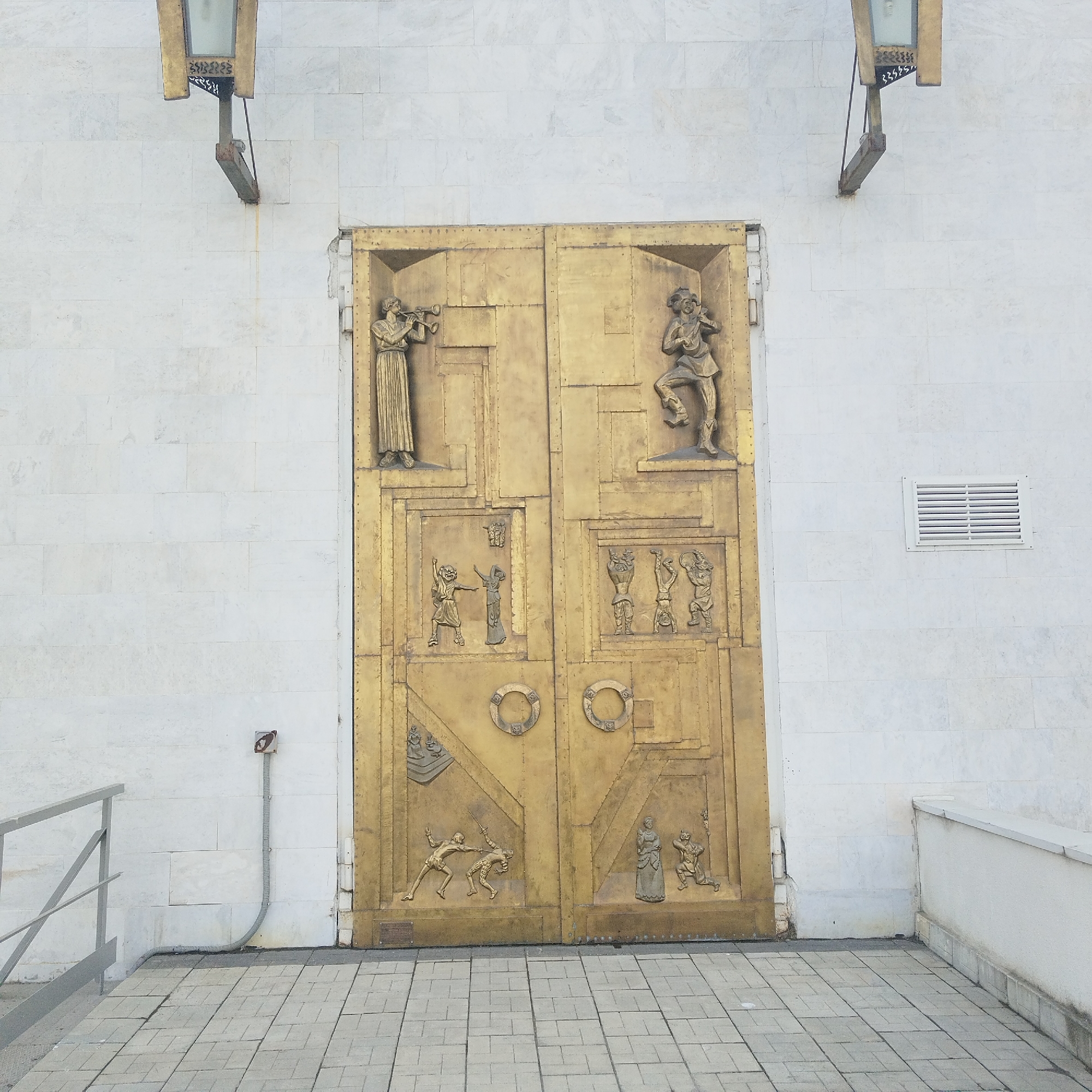 The image shows the entrance to a building, featuring a large, ornate golden door with multiple bas-relief panels depicting various scenes with human figures. Two vertical light fixtures are mounted on the building's exterior, flanking the door. The building's wall is made of light-colored, possibly marble, blocks. A gray tiled walkway leads up to the door, with a section of metal railing visible on the left. A vent is present on the wall to the right of the door. A conduit is attached to the wall on the left of the door. The photograph was taken in Yekaterinburg, Russia.