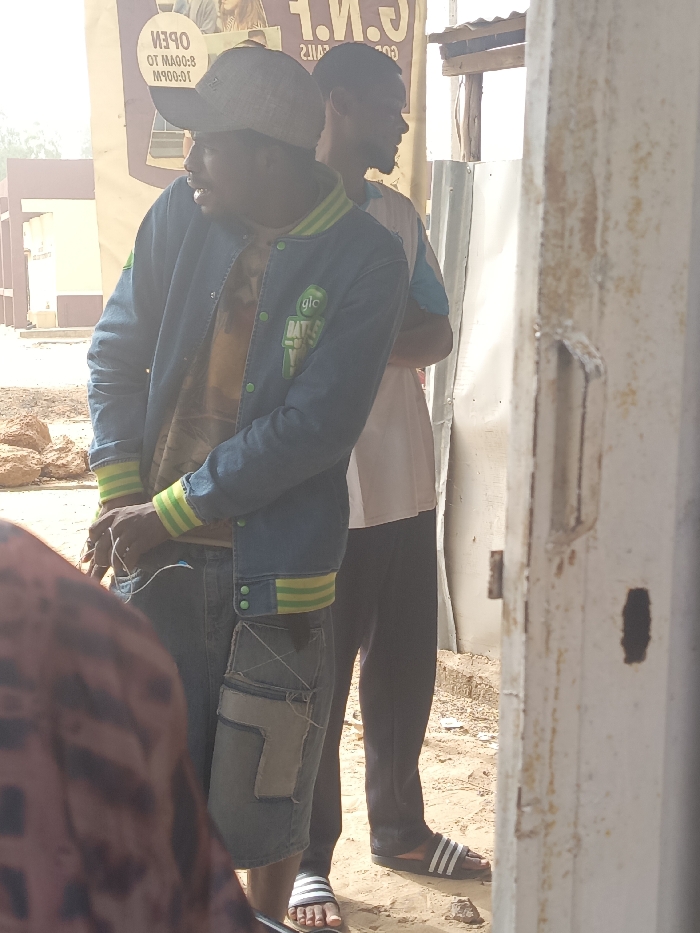 An outdoor scene in Kanti, Nigeria, shows three individuals in daylight. In the foreground, partially visible on the left, is the back of a person wearing a patterned reddish-brown garment. The central figure is a dark-skinned male, positioned slightly right of center, looking to his left. He wears a light-colored baseball cap, a blue denim jacket with green/yellow striped cuffs and collar, a multi-colored t-shirt underneath, and blue denim shorts with large, light-colored patch pockets on the thighs. White wires are visible in his hands, near his waist. He wears white and dark striped slide sandals. Behind him and to his right stands another dark-skinned male in profile, facing right. He wears a light-colored t-shirt with a blue accent, dark pants, and dark and white striped slide sandals, with his arms crossed. In the background, to the left, is a large, inverted banner advertising "G.N.F." and "OPEN 8:00 AM TO 10:00 PM." To the right, behind the second male, is a corrugated metal sheet structure and a basic wooden frame. The extreme right foreground is occupied by a heavily rusted, light-colored metal door frame or pillar featuring a handle and a rectangular aperture. The ground is unpaved, consisting of reddish-brown dirt and small stones. Distant, blurred reddish-brown buildings are visible in the far background on the left.