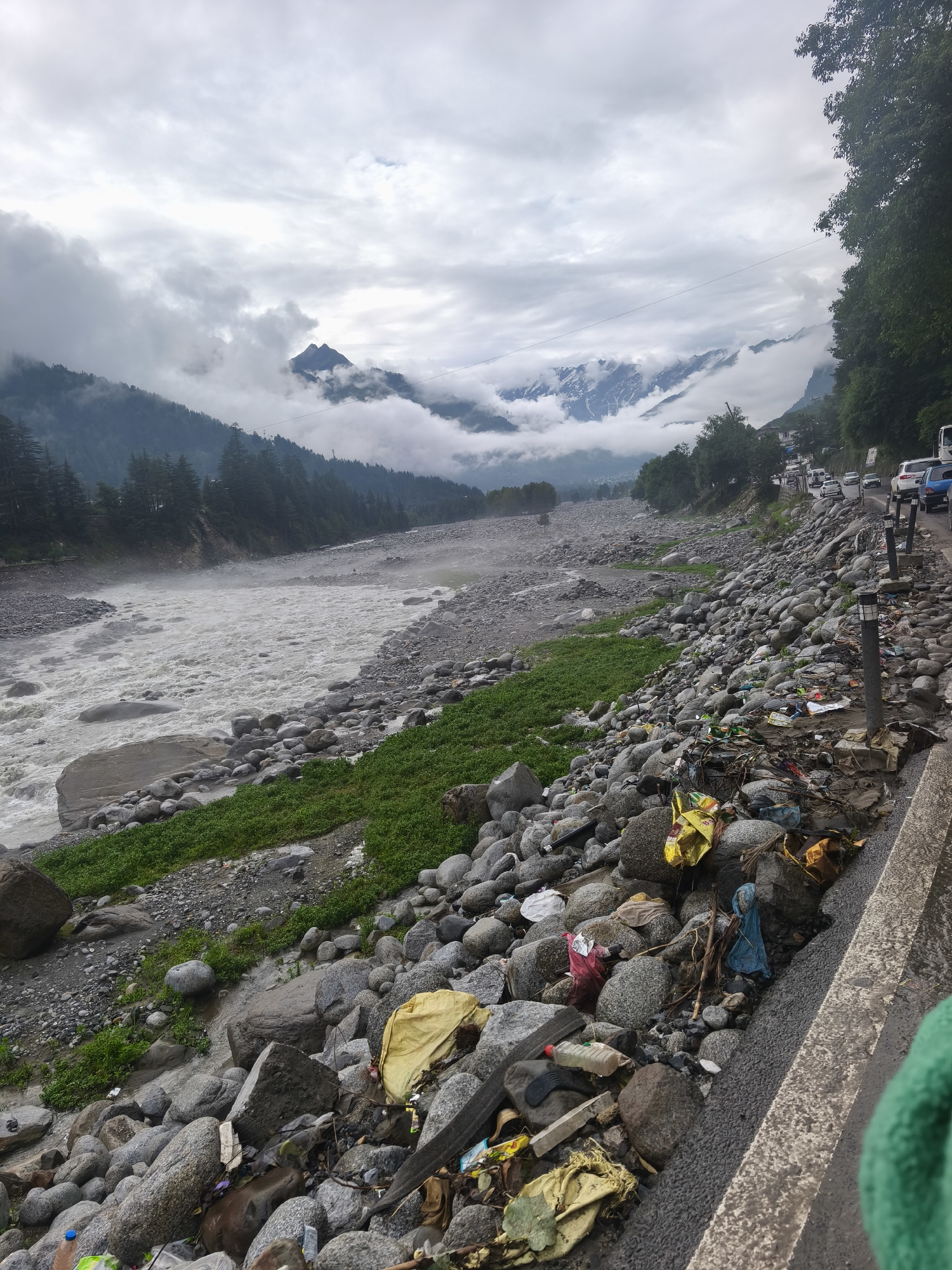 The image captures a scenic mountain landscape in Vashisht, India, under a cloudy sky. A wide, turbulent river flows through a rocky valley, its waters appearing murky and fast-moving. The riverbanks are strewn with large grey stones and patches of vibrant green grass.
On the right side of the image, a road runs alongside the river. This road is lined with a significant amount of scattered garbage, including plastic bags, bottles, and other debris, creating a stark contrast with the natural beauty of the surroundings. Several streetlights are positioned along the edge of the road, suggesting it is a developed area. In the distance, a traffic jam is visible on the road, with numerous cars and buses lined up.
The mountains in the background are partially obscured by thick clouds, adding a dramatic and somewhat melancholic atmosphere to the scene. The time of day appears to be daytime, given the overall illumination, but the overcast conditions suggest it might be a gloomy or pre-rainy day. The presence of traffic and the scattered garbage indicate human activity and its impact on the environment in this otherwise breathtaking location. The image evokes a sense of both natural grandeur and the unfortunate reality of pollution. No visible text is present in the image.