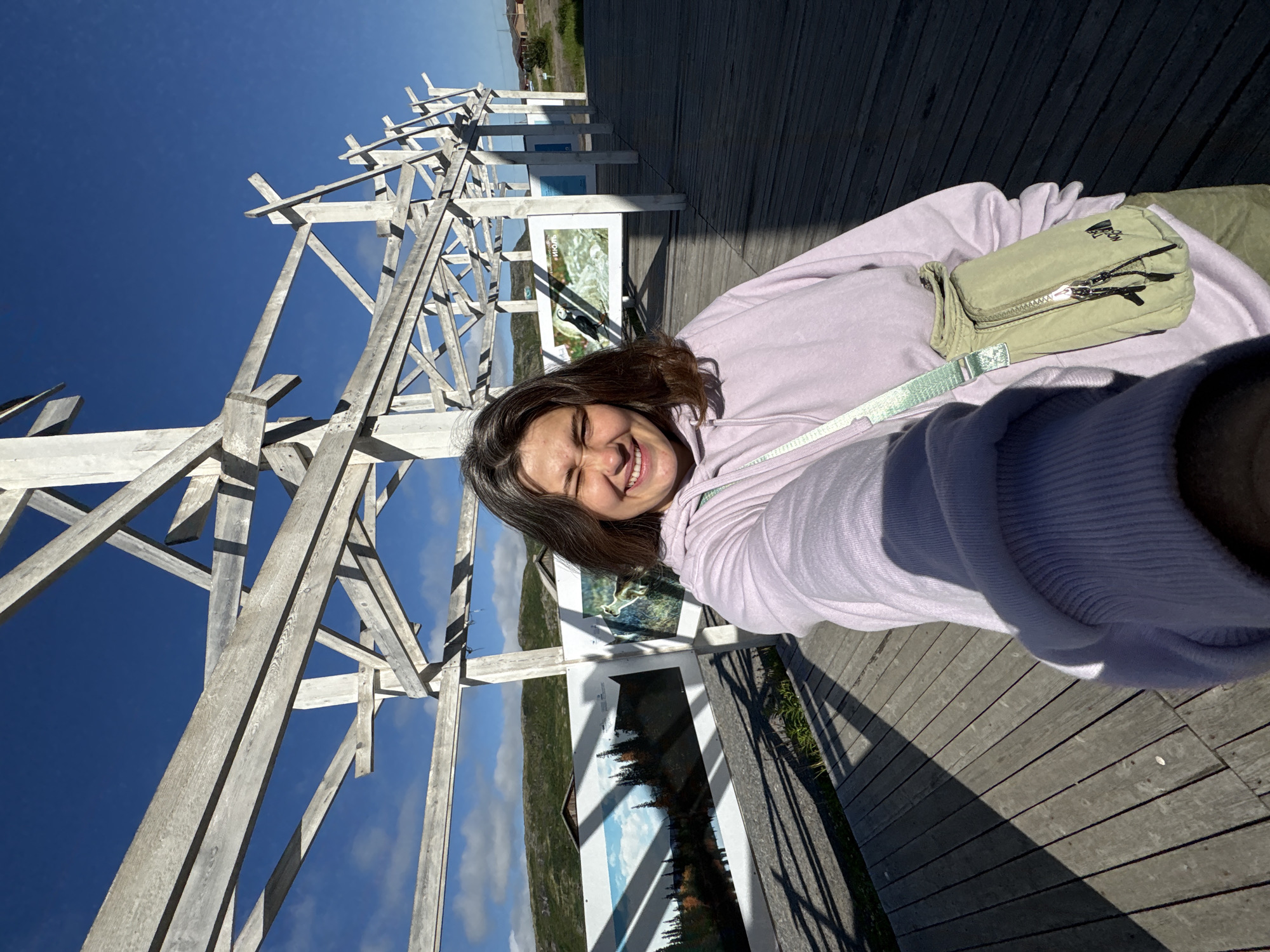 In Teriberka, Russia, a young woman with shoulder-length brown hair smiles for a selfie in front of an outdoor art exhibition. She wears a light purple hoodie and a light green crossbody bag. The exhibition features large landscape photographs mounted on white panels, displayed along a wooden boardwalk. A skeletal wooden structure, possibly an unfinished pavilion or an art installation itself, arches overhead. The scene is bathed in bright sunlight, casting distinct shadows on the wooden planks and the surrounding grassy landscape, which features rolling hills and distant buildings. The sky is a clear, vibrant blue.