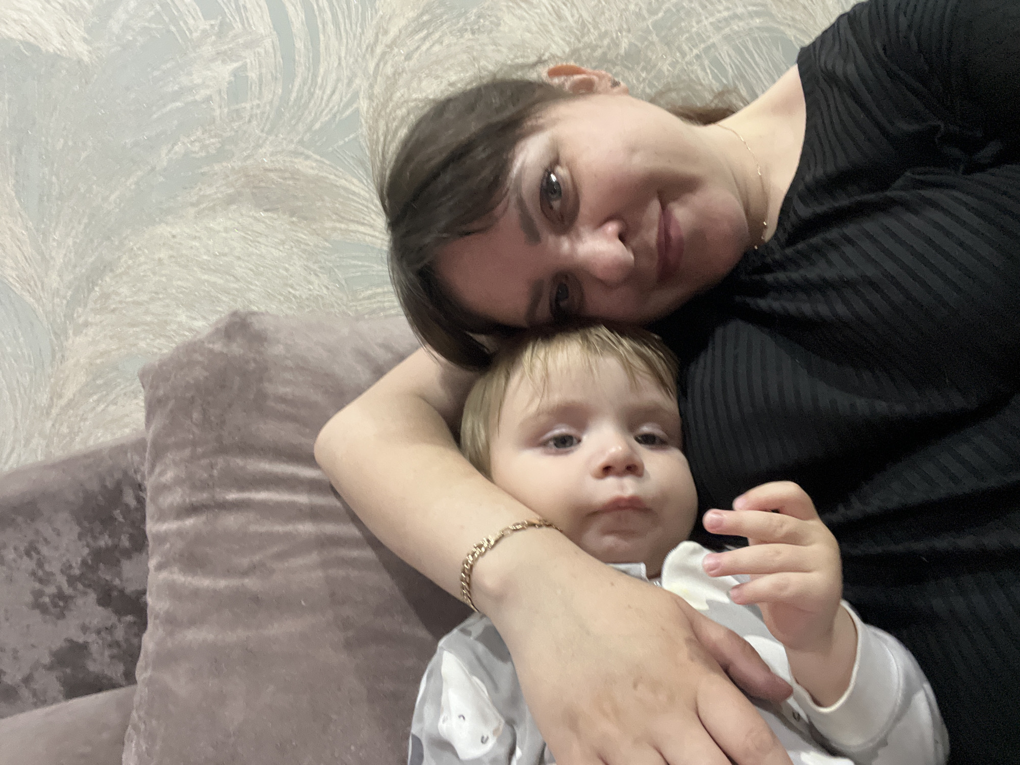 A woman and a child are lying down on a couch in a room. The woman is wearing a black shirt and has dark brown hair and brown eyes. She is looking at the camera with a slight smile. The child is wearing a grey and white patterned onesie. The woman's arm is around the child. The location and the city of Noviy, Russia cannot be determined from the image.