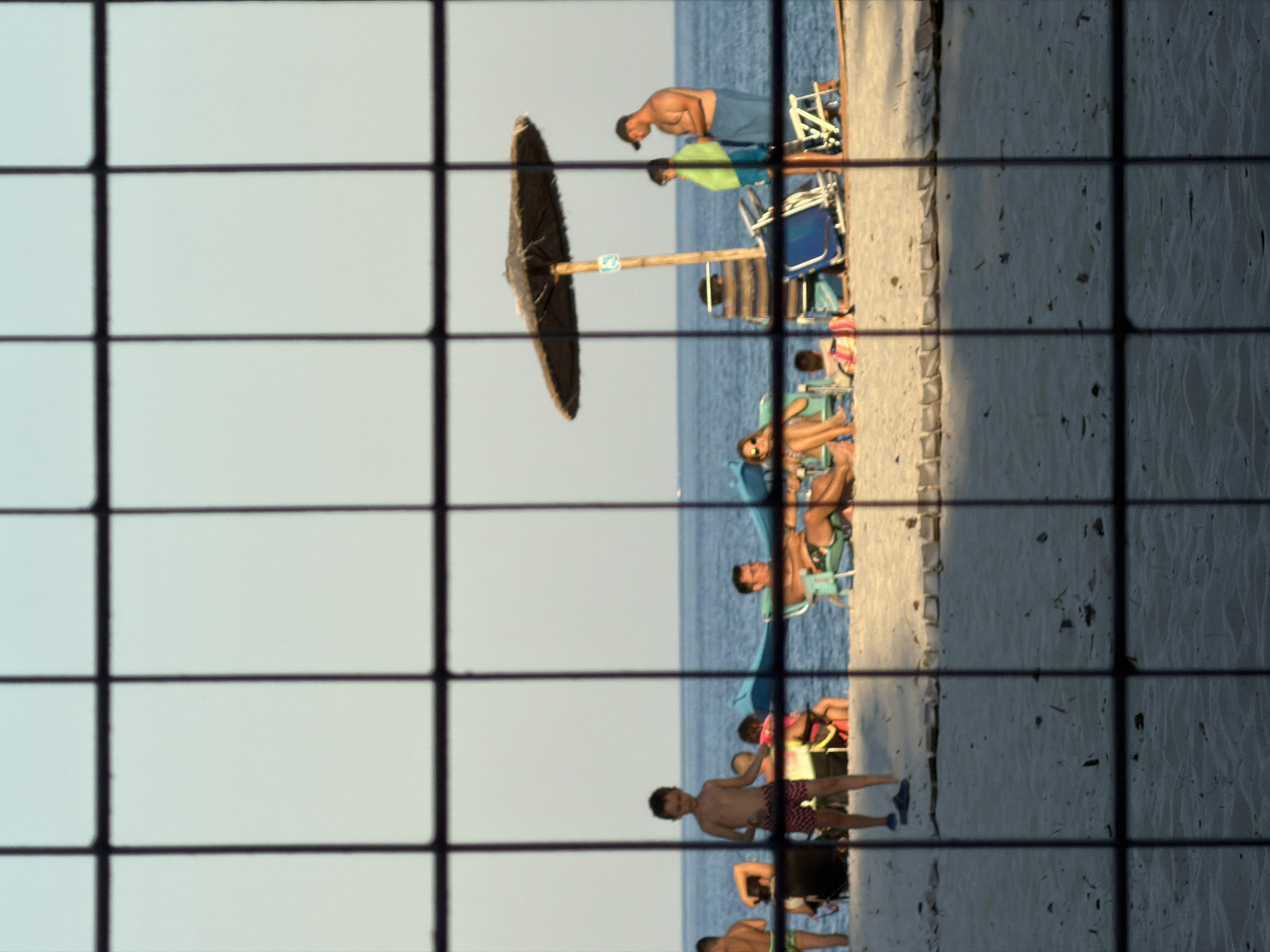 This is a medium shot, through a fence, of a beach scene at Golden Beach, Greece, during daylight. The foreground is dominated by the dark, grid-like structure of a fence, obscuring the lower half of the image.
In the mid-ground, a sandy beach stretches towards the sea. Several people are visible, some sitting on beach chairs, others standing. On the left, a young boy, wearing patterned shorts, stands on the sand, holding a bright pink object. To his right, other beachgoers are partially visible, some reclining in chairs. Further to the right, a straw umbrella provides shade for a beach area. Two people, a man and a woman, are standing near the umbrella, with the man appearing to be talking to the woman. Several beach chairs, some folded and some occupied, are scattered on the sand. The sea is a calm expanse of blue water, meeting a pale, cloudless sky. The overall atmosphere is relaxed and suggests a typical summer day at the beach.