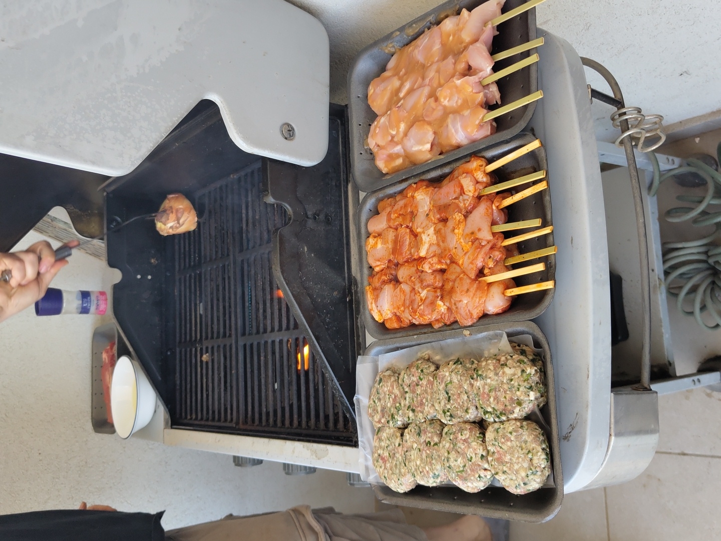 The image depicts a barbecue grill with a single piece of meat cooking on the grates. To the right of the grill are three trays of uncooked food: chicken skewers, marinated chicken skewers, and hamburger patties. A person is visible in the bottom center of the frame, and another person is visible using a grilling utensil near the grill. The location cannot be determined from the image.
