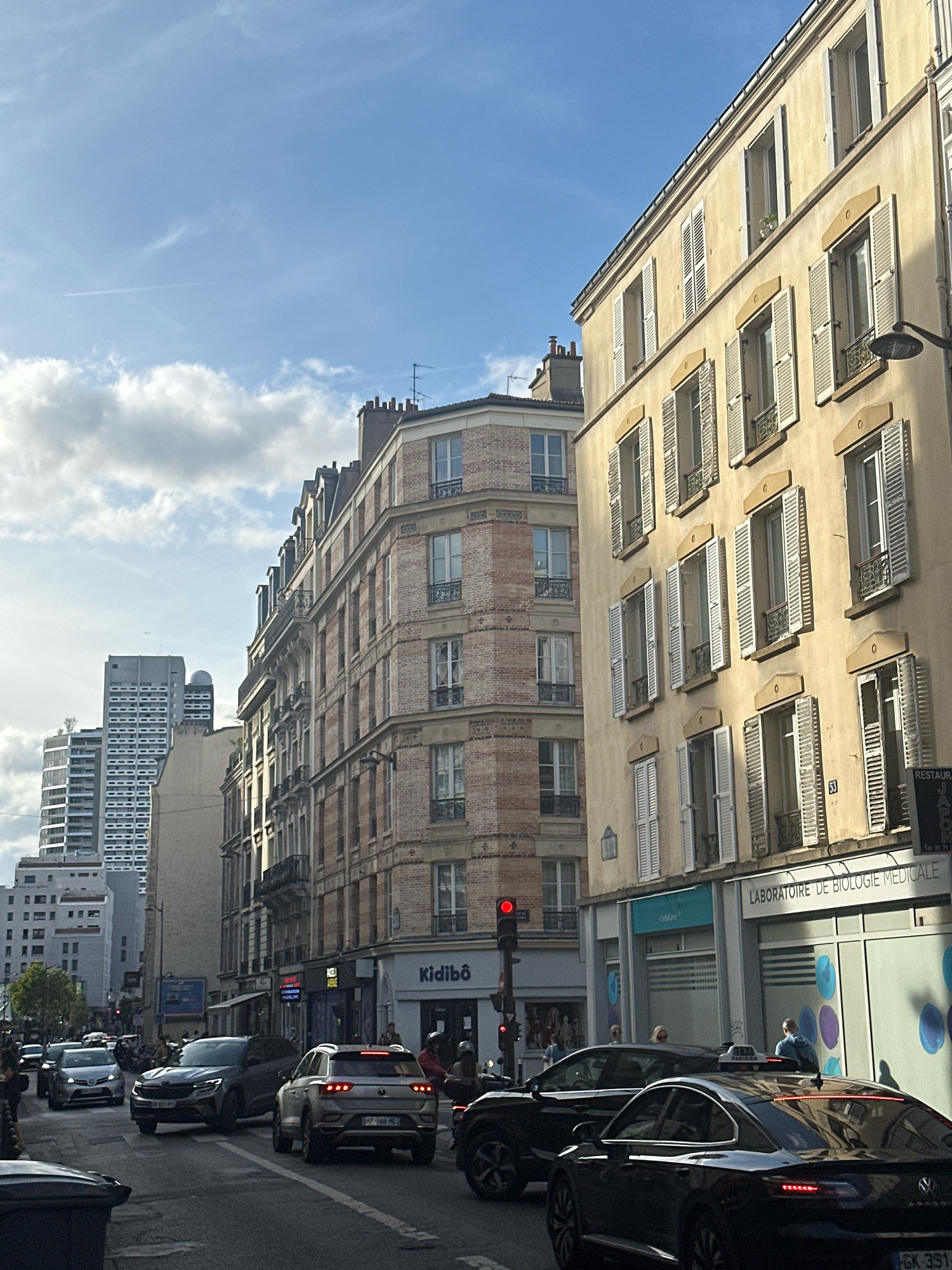 The image depicts a street scene in Paris, France, with a prominent building on the right side of the frame. The building is a light yellow facade with multiple floors, each featuring numerous windows with white shutters, some open and some closed. The architecture suggests a classic Parisian style. In front of the building, there is a storefront with a sign that reads "LABORATOIRE DE BIOLOGIE MÉDICALE." Another smaller sign above it says "Kidibo." A traffic light is visible in the middle of the street, showing a red signal. Several cars are on the road, indicating moderate traffic. Some pedestrians are visible on the sidewalk and in the street, including a person wearing a blue backpack. In the background, taller, modern buildings can be seen, contrasting with the older architecture. The sky is partly cloudy with a clear blue section visible. The overall scene is a typical urban street view in Paris, capturing a moment of daily life.