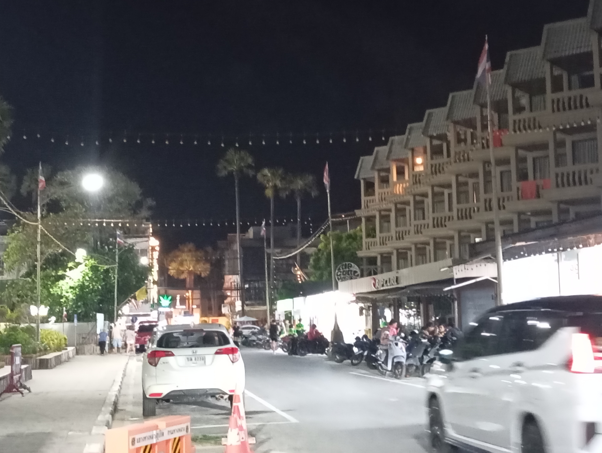 The image shows a street scene at night in Karon, Thailand. On the right, a multi-story building with balconies and a tiled roof is illuminated. A Thai flag flies from a flagpole in front of the building. In the foreground, a white car is parked on the left side of the street. Further down the street, several motorbikes are parked and some people are gathered. The street is lined with trees and some businesses with illuminated signs. The overall impression is a busy street at night, likely in a tourist area.
