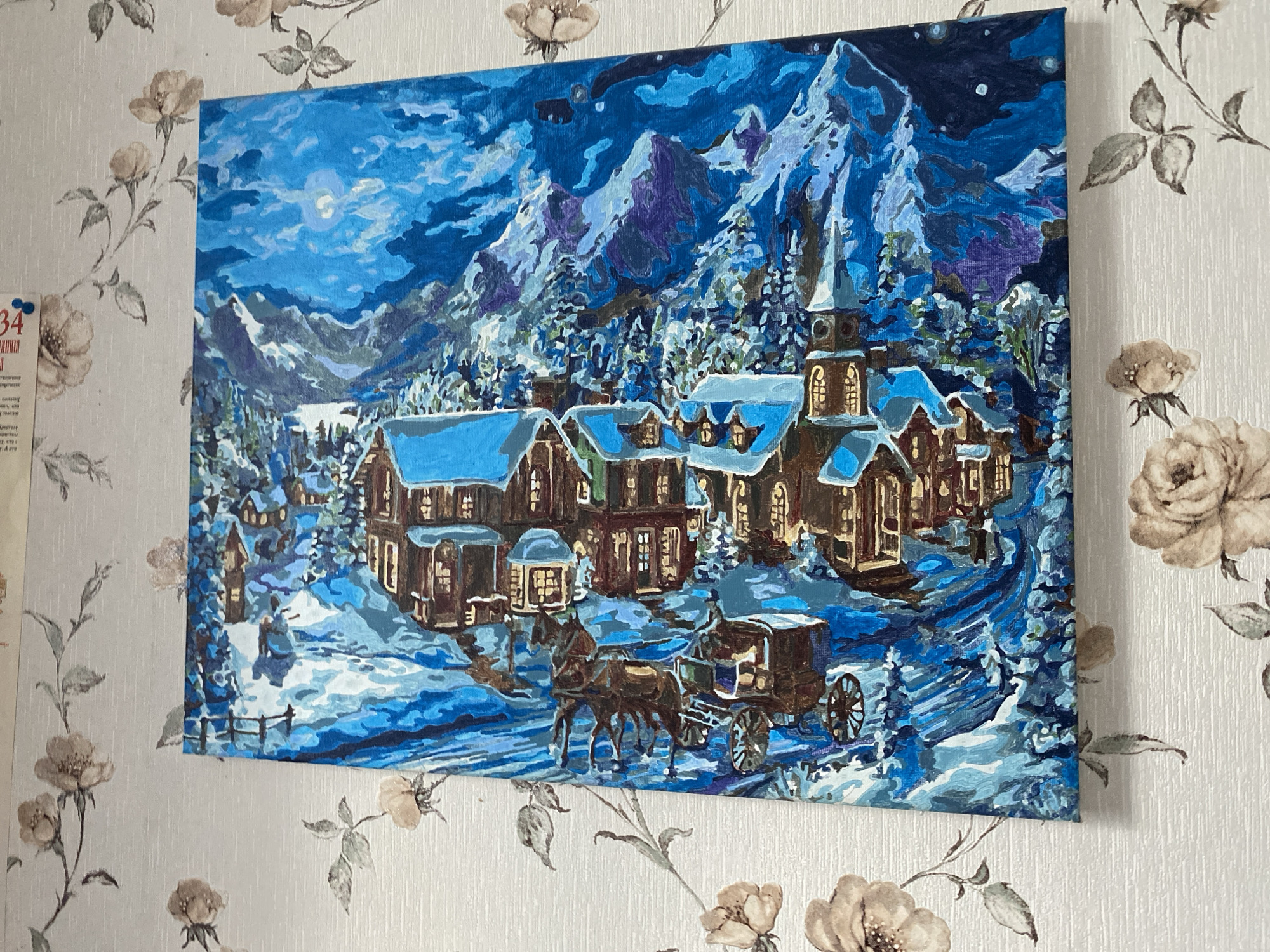 The image shows a painting of a winter village scene. In the foreground, a horse-drawn carriage is moving through a snow-covered path. Behind the carriage, there are several houses with lit windows, suggesting warmth and life within. A prominent building with a steeple, resembling a church or town hall, stands in the center. In the background, snow-laden trees and majestic mountains create a picturesque backdrop under a bright, cloudy moonlit sky. The painting is hung on a wallpapered wall adorned with a floral pattern. The village appears to be located in Vvedenka, Ukraine, as indicated by external information not present in the image itself.