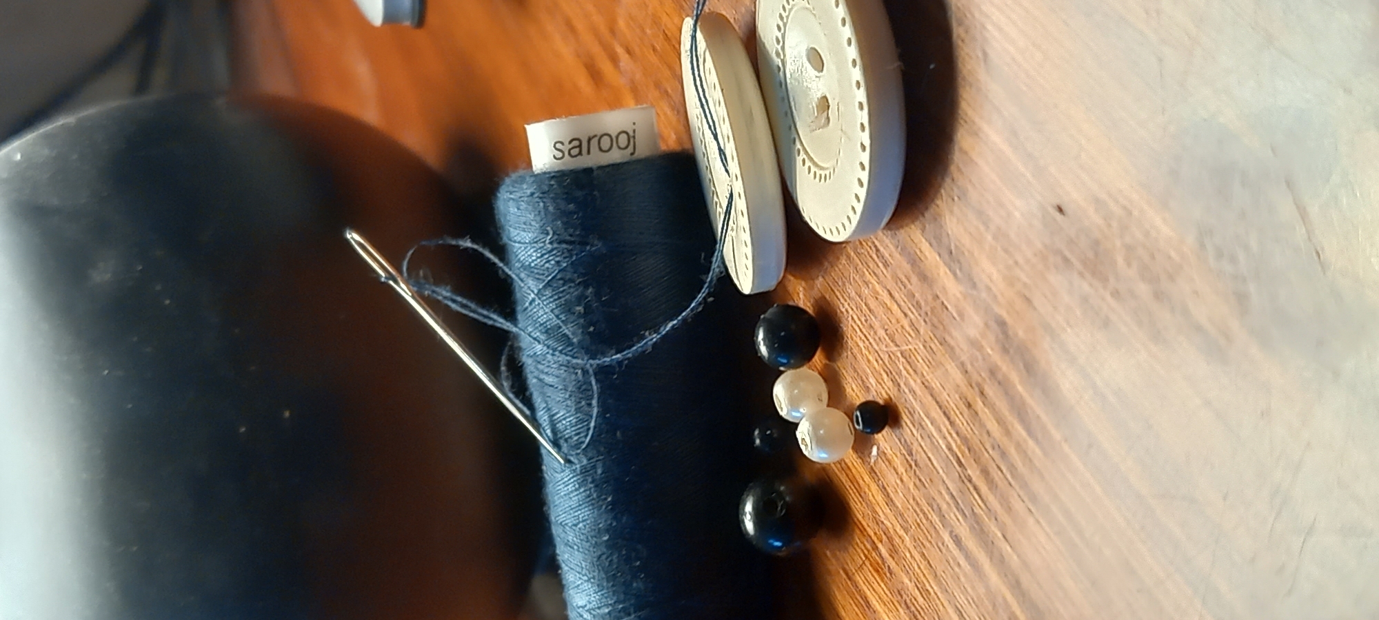 The image displays a collection of sewing and crafting materials arranged on a polished wooden surface, likely a table or desk, in an indoor setting.
The central subject is a spool of dark blue thread, from which a silver sewing needle protrudes, threaded with a loop of the same blue thread. A small, white rectangular label is visible on top of the thread spool, clearly showing the text "sarooj".
To the right of the thread spool, there are two large, round, cream-colored buttons. Both buttons feature a decorative edge with numerous small, uniform holes or indentations, and a central hole for attachment. One button lies flat, while the other is slightly angled. Adjacent to the buttons and thread, a small cluster of beads is scattered. These beads come in two main colors: glossy black and iridescent white or cream, varying slightly in size.
The wooden surface exhibits a noticeable grain pattern and reflects some light, indicating a smooth finish. In the background, particularly on the left side, a dark, indistinct object, possibly a container or part of furniture, is visible and out of focus. The lighting appears consistent with an indoor environment, potentially from a window or an overhead light source, creating subtle highlights and shadows. No active event is captured; the items appear to be neatly laid out, suggesting either a paused craft project or an organized collection of supplies. The scene is located in Kryvyi Rih, Ukraine.