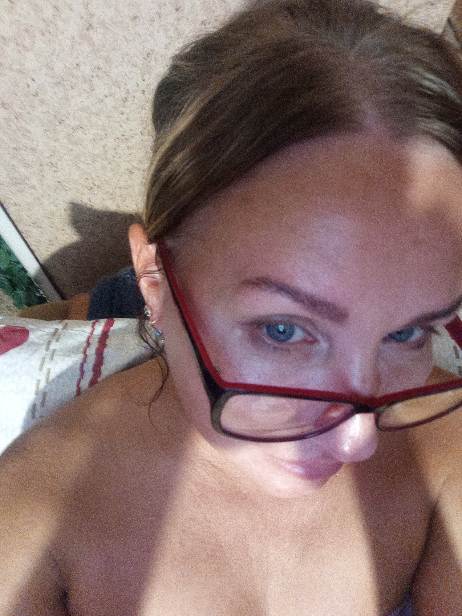 A close-up, overhead, low-angle, indoor shot captures a person from the shoulders up. The individual is wearing red-rimmed glasses and has light brown hair pulled back. Their eyes are a striking blue. The background consists of a textured, light-colored wall and a striped pillow with red and white stripes, and a red heart detail. The lighting creates a strong shadow across the person's chest and shoulder. The overall scene suggests a casual, personal moment within a home environment. The location is not discernable from the image.