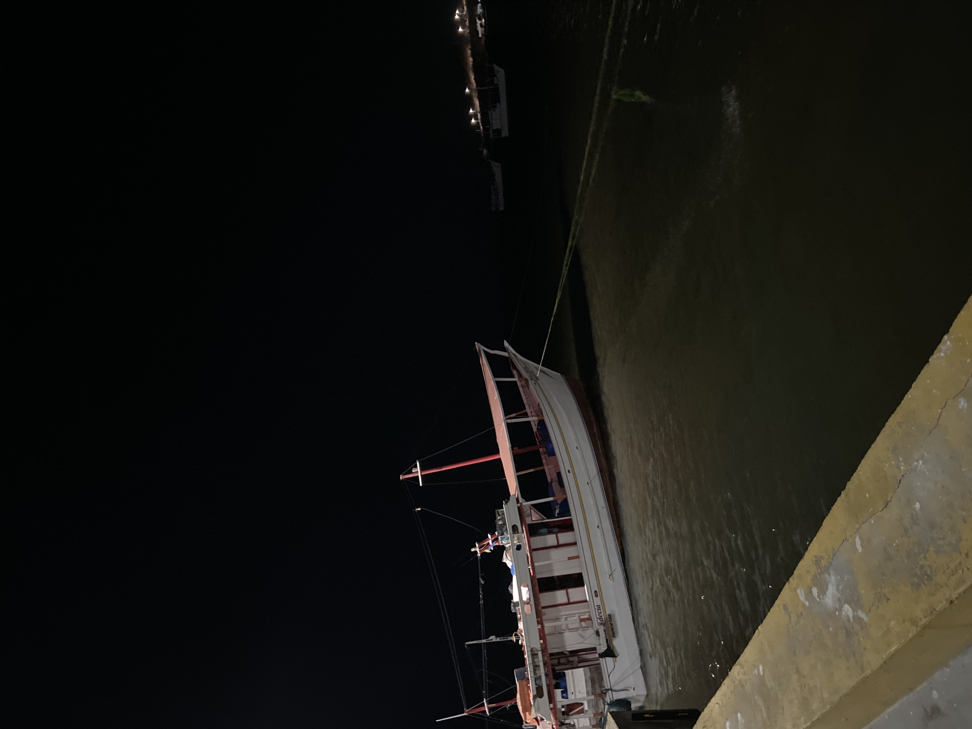 At night, a medium-sized white boat with red trim is docked alongside a concrete pier in Juan Griego, Venezuela. The water appears dark and murky, with other boats visible in the distance. The sky is dark, and the overall lighting suggests a coastal setting. The boat is secured with a rope to the dock. The word "Julecia" is visibly written on the side of the ship. The lighting is poor, but sufficient to discern the features of the boat and the surrounding dock.