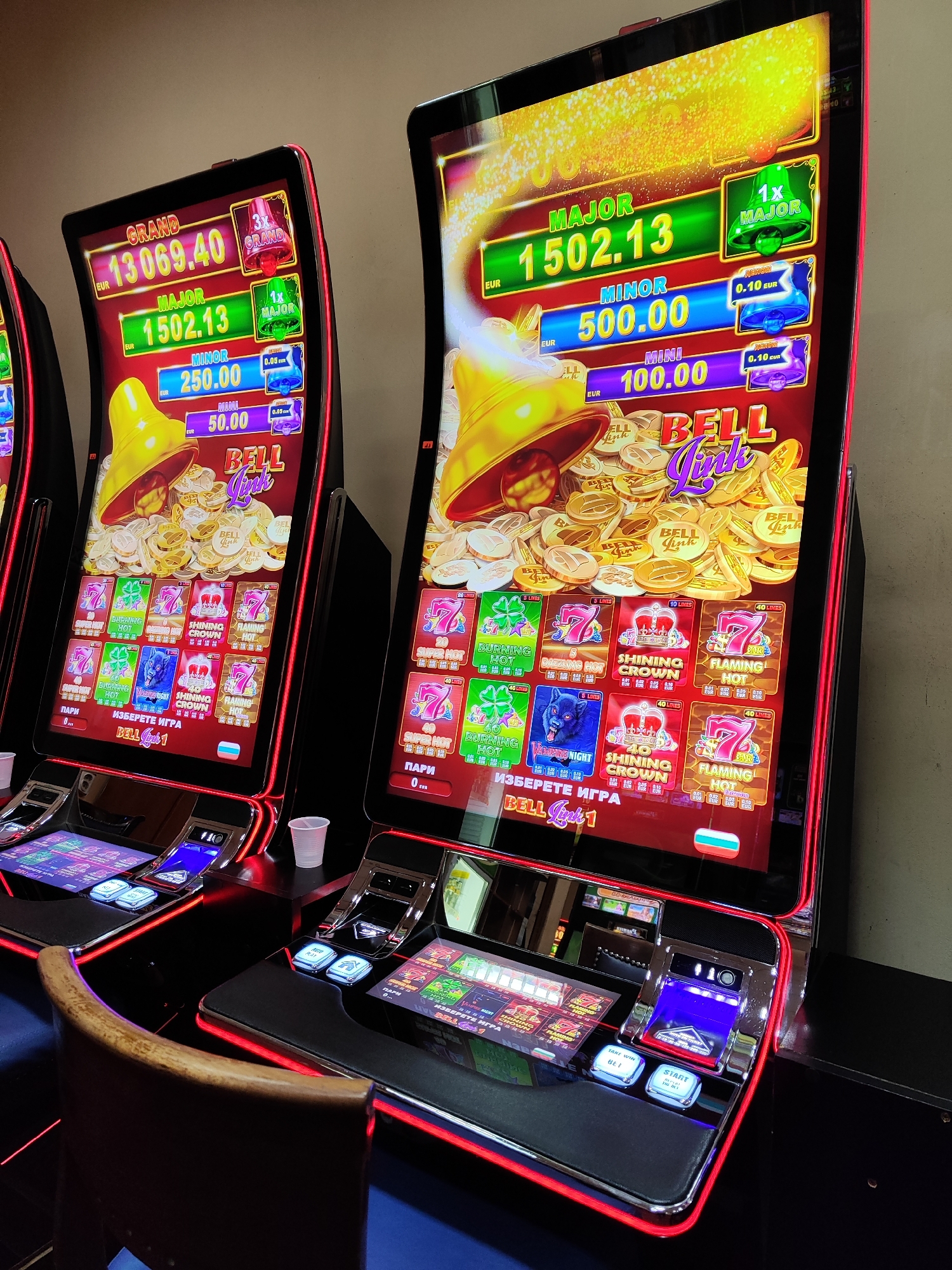 The image shows two "Bell Link" slot machines, possibly located in a casino or gambling establishment. The machines have curved screens displaying a golden bell and coins with "Bell Link" written on them, along with various game options such as "Burning Hot," "Shining Crown," and "Flaming Hot." The left machine shows a "Grand" prize of 13069.40 EUR and a "Major" prize of 1502.13 EUR, while the right machine shows a "Major" prize of 1502.13 EUR, a "Minor" prize of 500.00 EUR, and a "Mini" prize of 100.00 EUR. Both machines have buttons and interfaces for placing bets and starting the game, with a cup visible near one machine. The machines are dark in color with red LED highlights, and a wooden chair is partially visible near the left machine. A third machine is partially visible to the left.