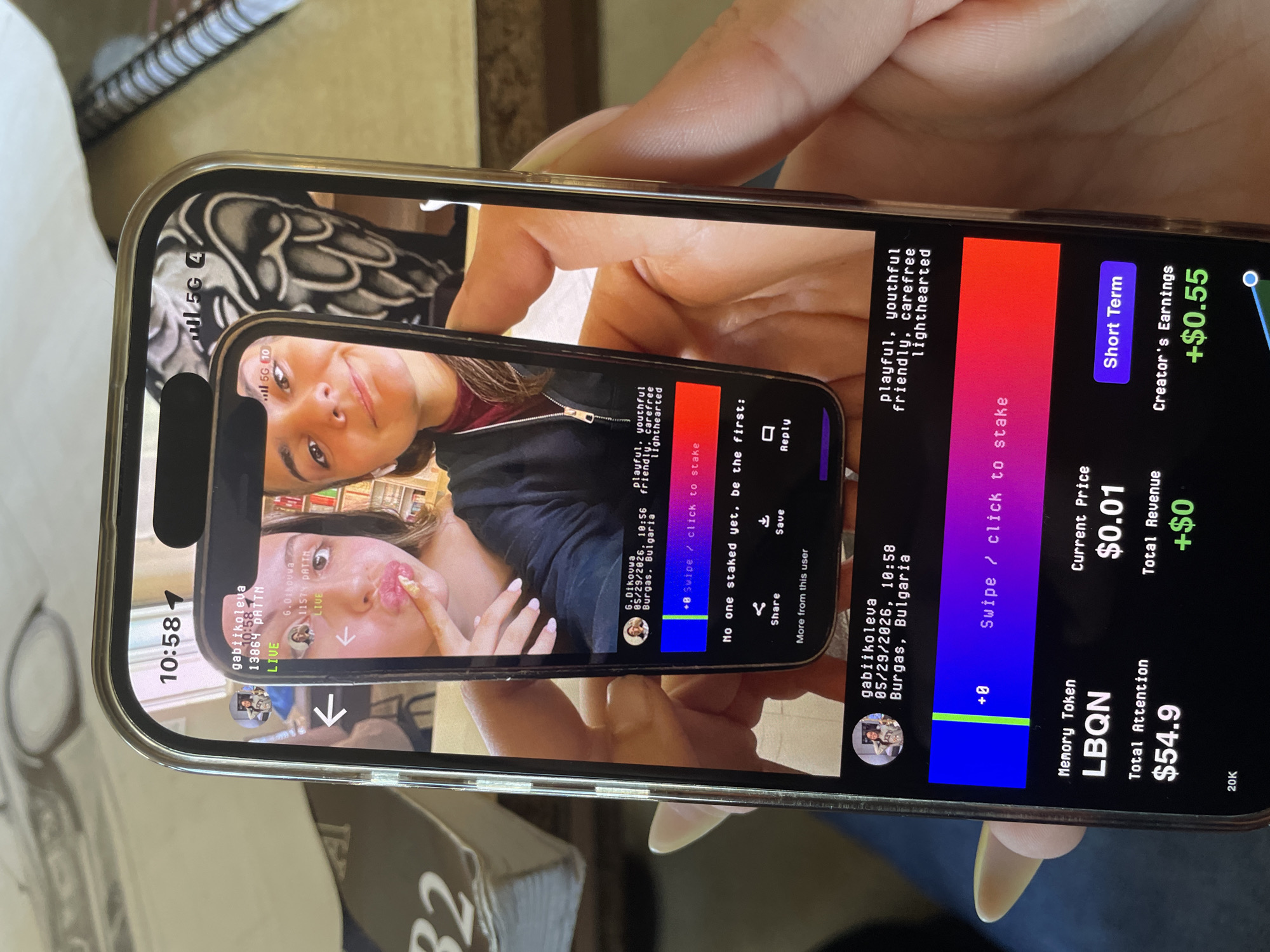 A person is holding a smartphone displaying a live video stream on what appears to be a social media platform. The video features a young woman with long brown hair, seemingly interacting with the viewer, as evidenced by her gestures and facial expressions. The interface on the smartphone indicates it's a live broadcast ("LIVE") from gabiikoleva, a user in Burgas, Bulgaria. The overlaid text specifies a date of "05/29/2026, 10:56" suggesting a future broadcast date. The app shows details like "Current Price $0.01", "Total Attention $54.9," and "Creator's Earnings +$0.55," indicating a potential monetization feature. The surrounding environment consists of a table or desk with a notebook. The location mentioned in the phone display is Burgas, Bulgaria.